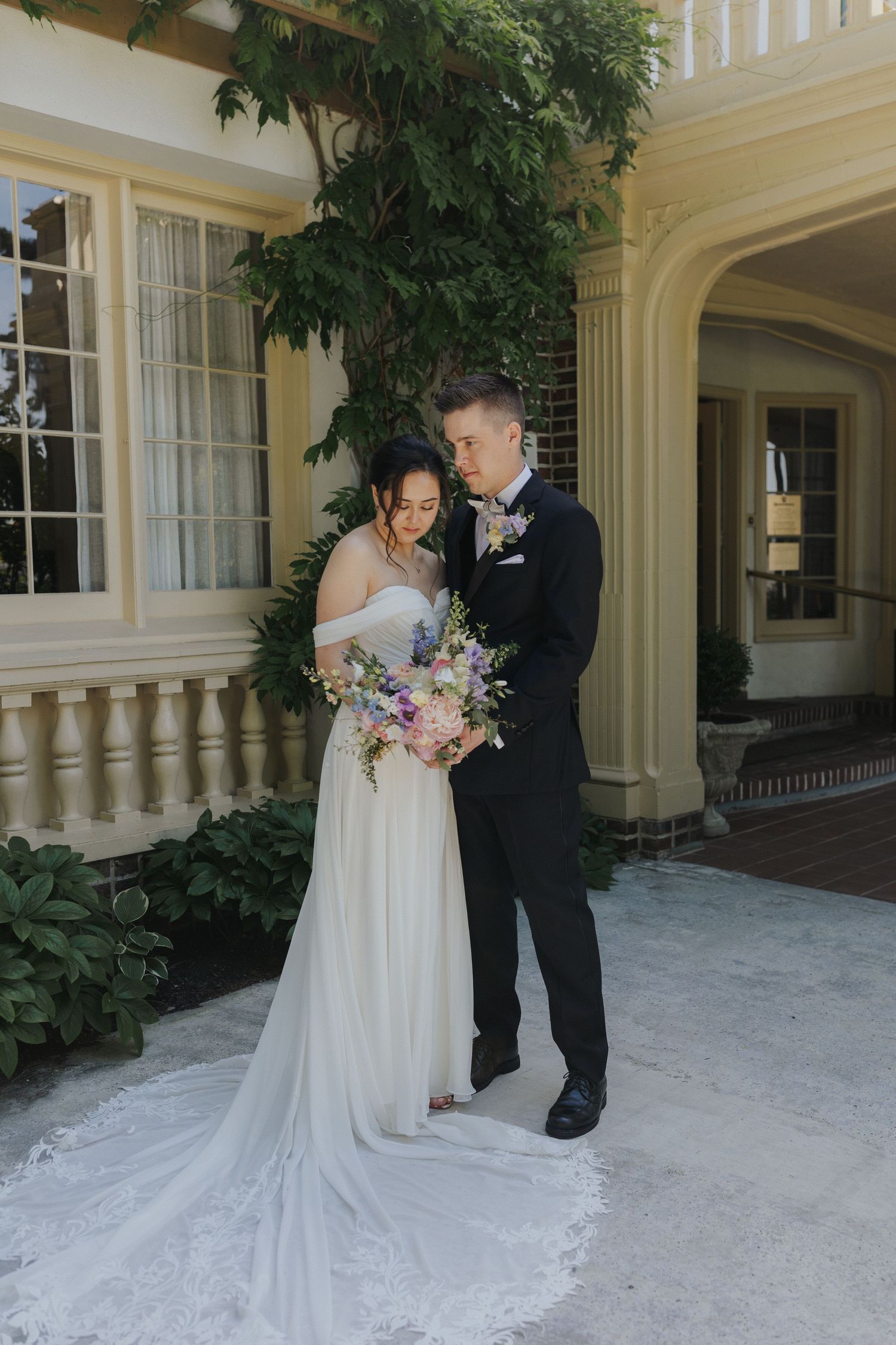Elegant wedding portrait under mansion archway with lush greenery and floral bouquet.