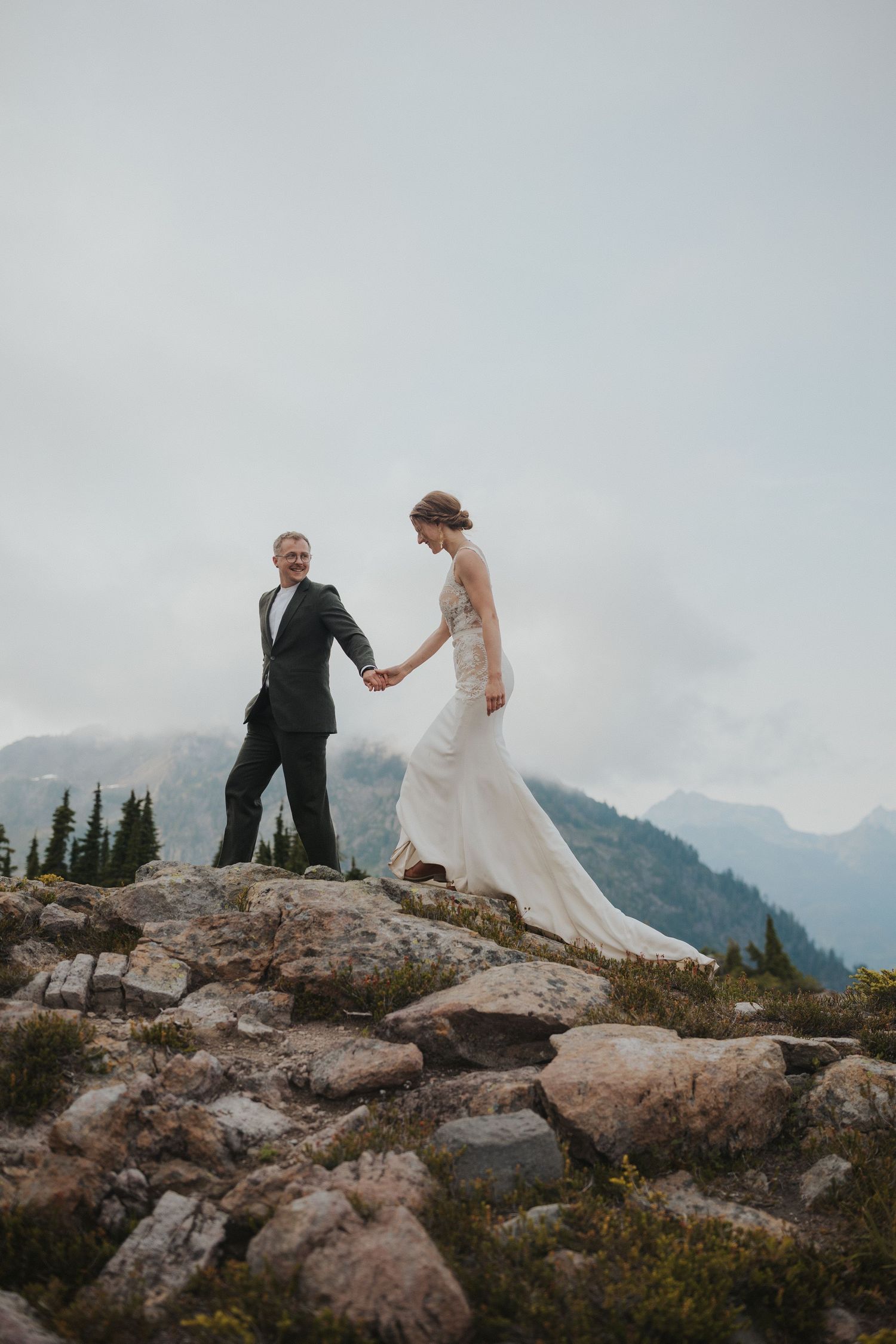 Two people in wedding attire explore a rocky mountain summit with misty peaks in the distance.