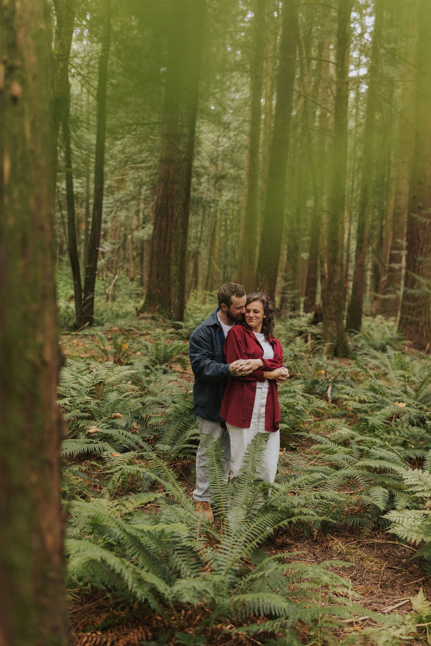 A couple embraces among tall trees and ferns in a lush forest setting.