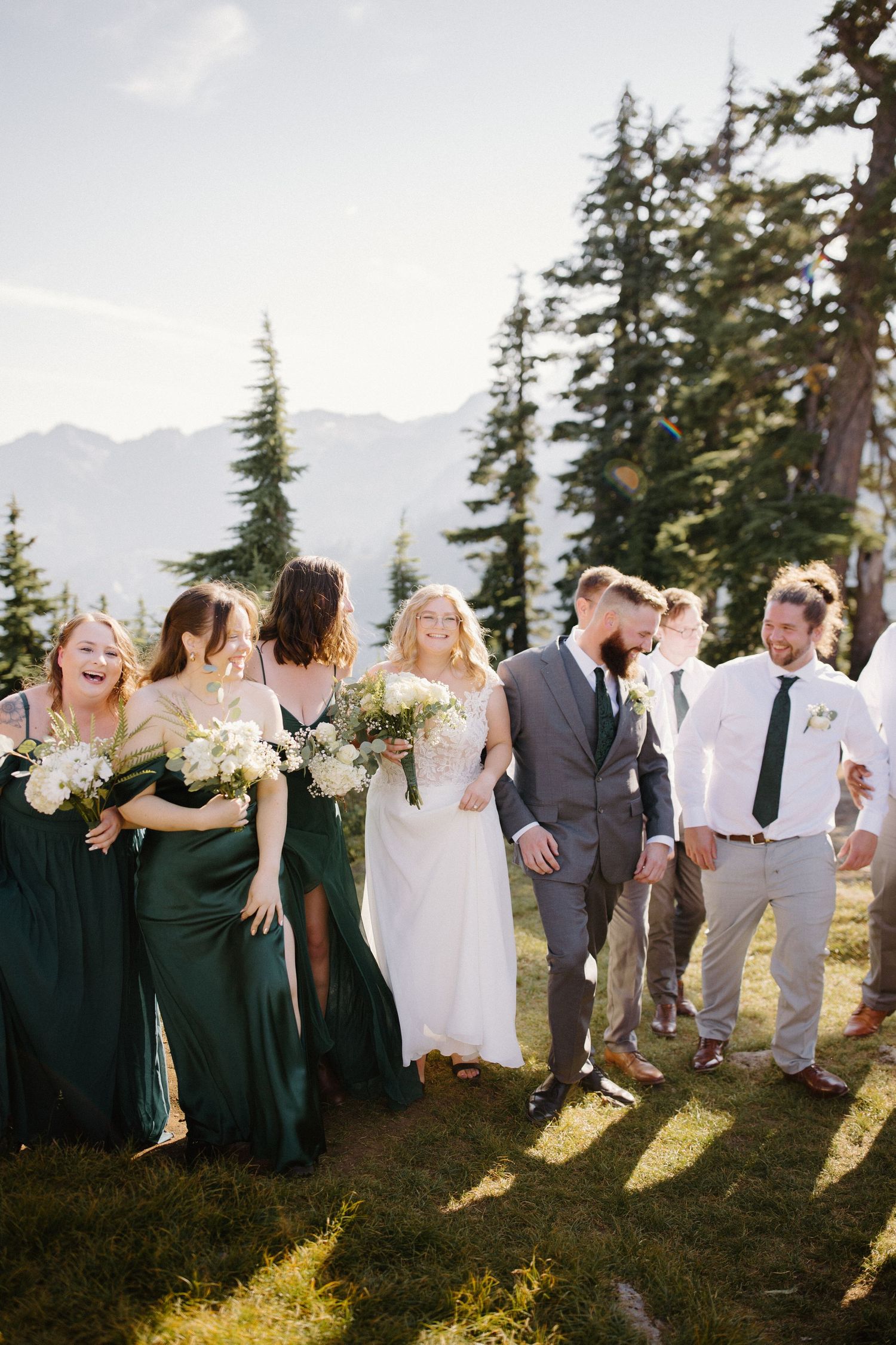 Wedding party laughs together on a sunny mountain slope with dramatic mountain views.