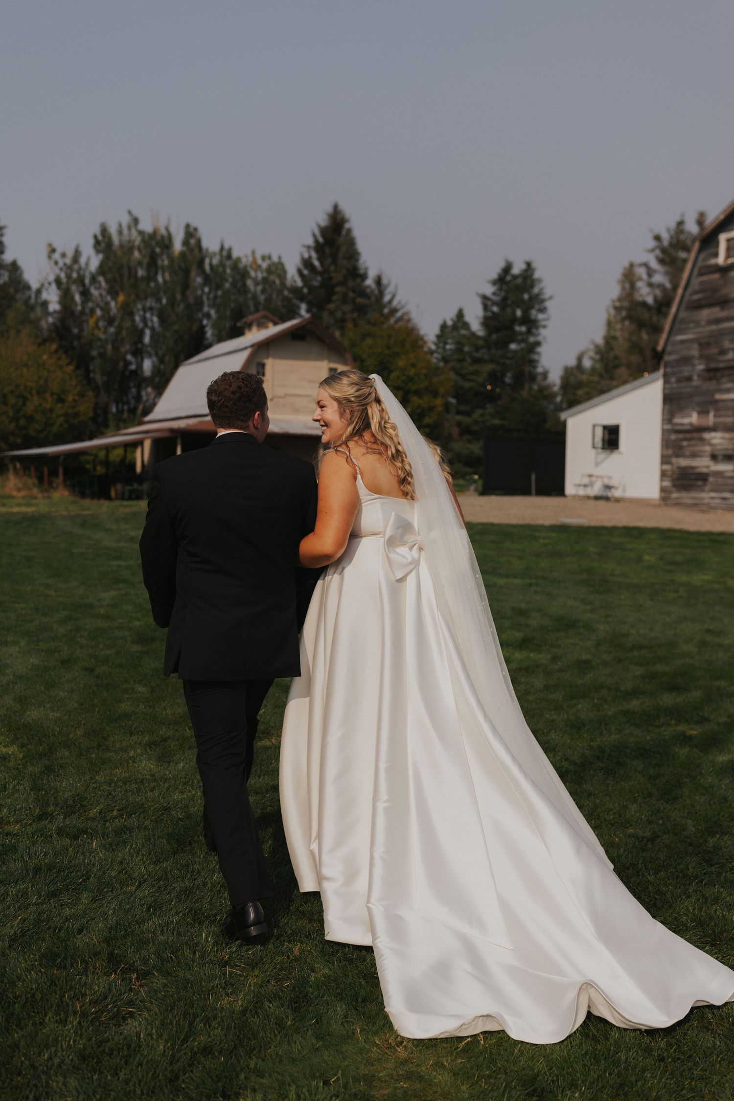 A bride in a flowing white gown walks through a grassy field during golden hour.