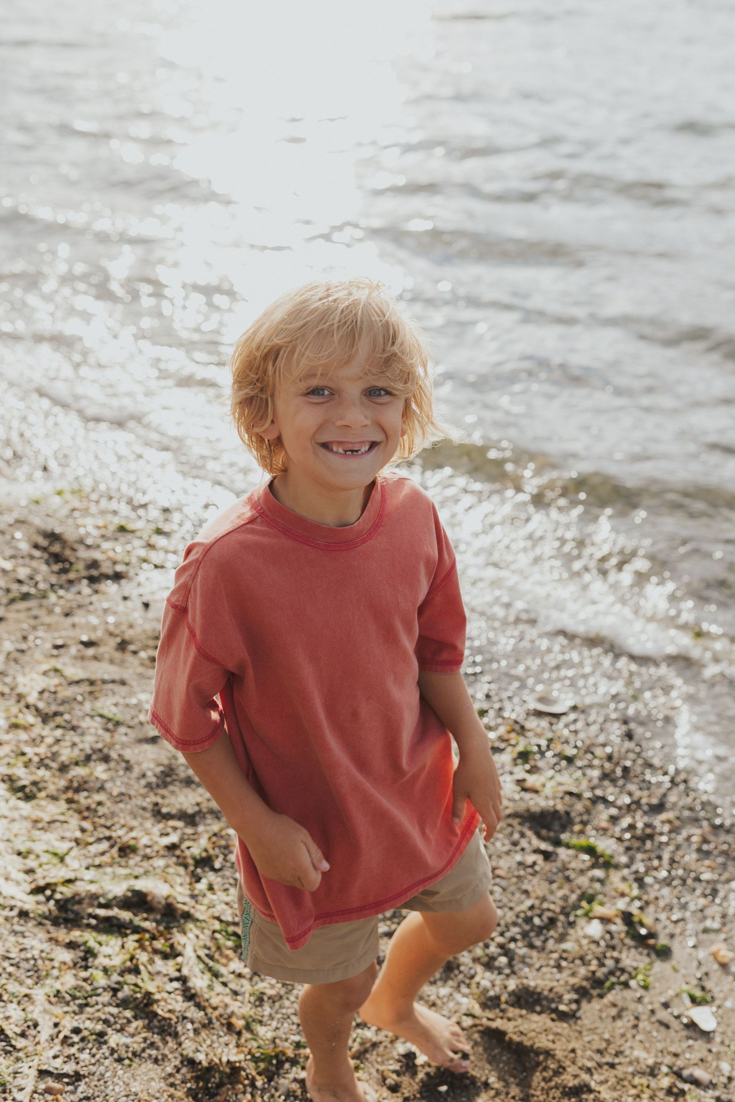 Child in red shirt playing and smiling on rocky beach near the water.