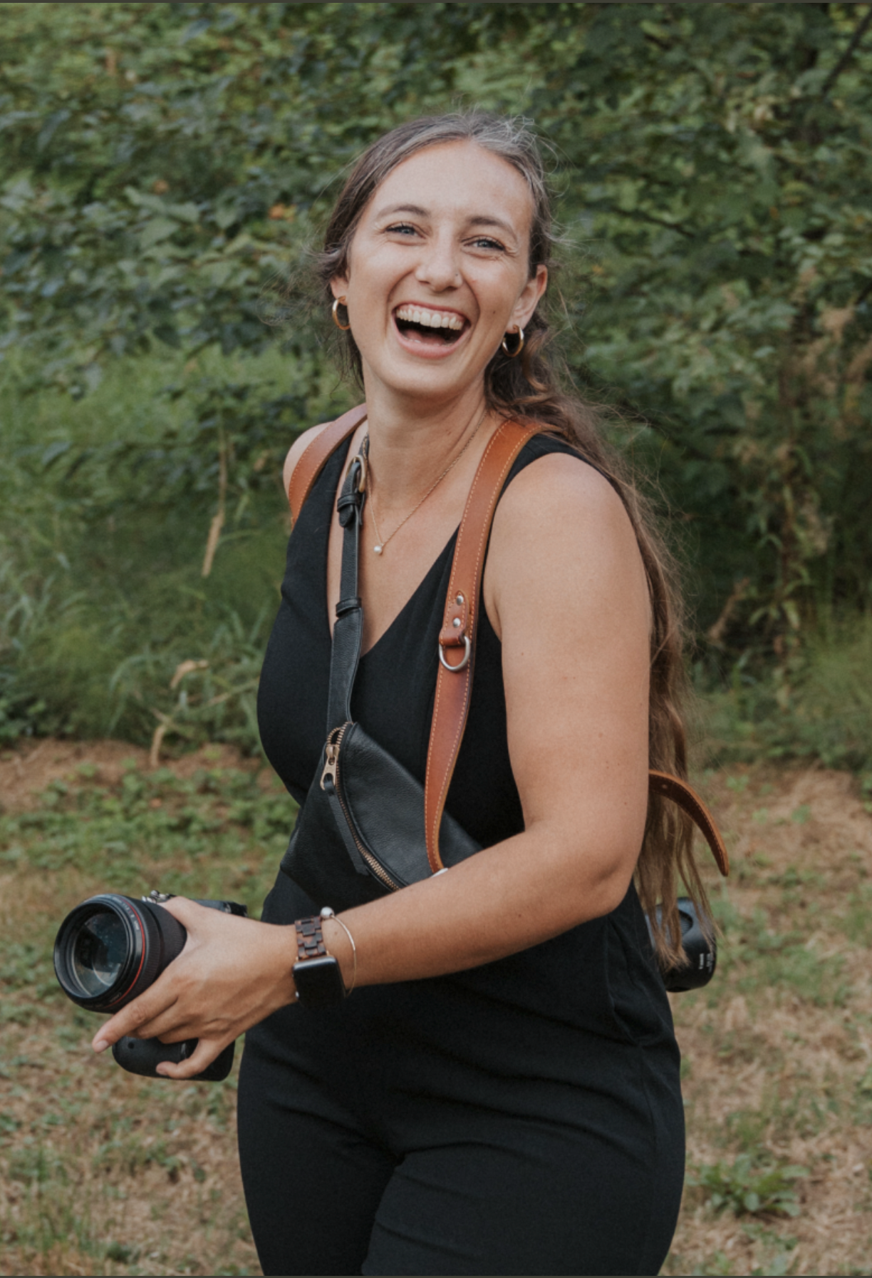 Photographer in black dress laughing joyfully while holding camera equipment outdoors.