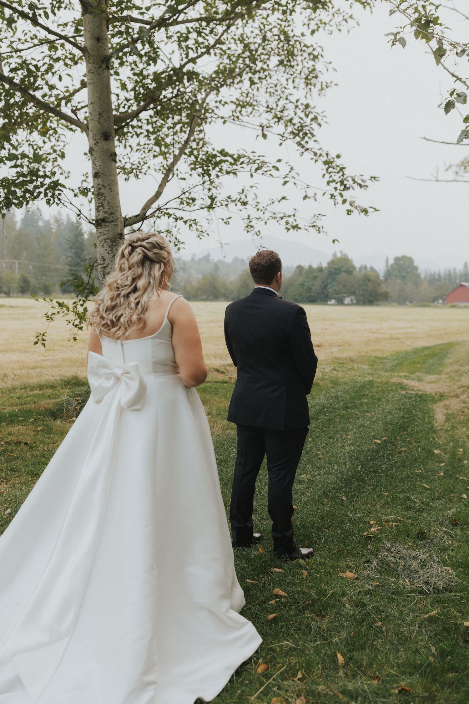 A couple in wedding attire stands together on a grassy hillside surrounded by trees.