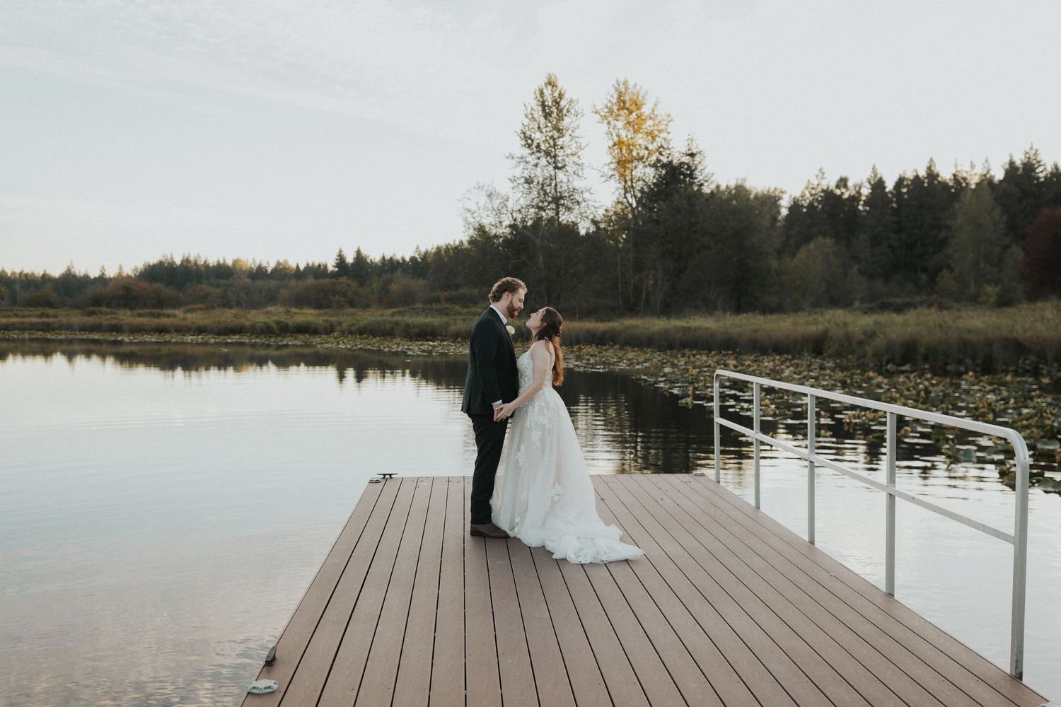 Wedding couple embracing on wooden dock overlooking serene lake and forest landscape.