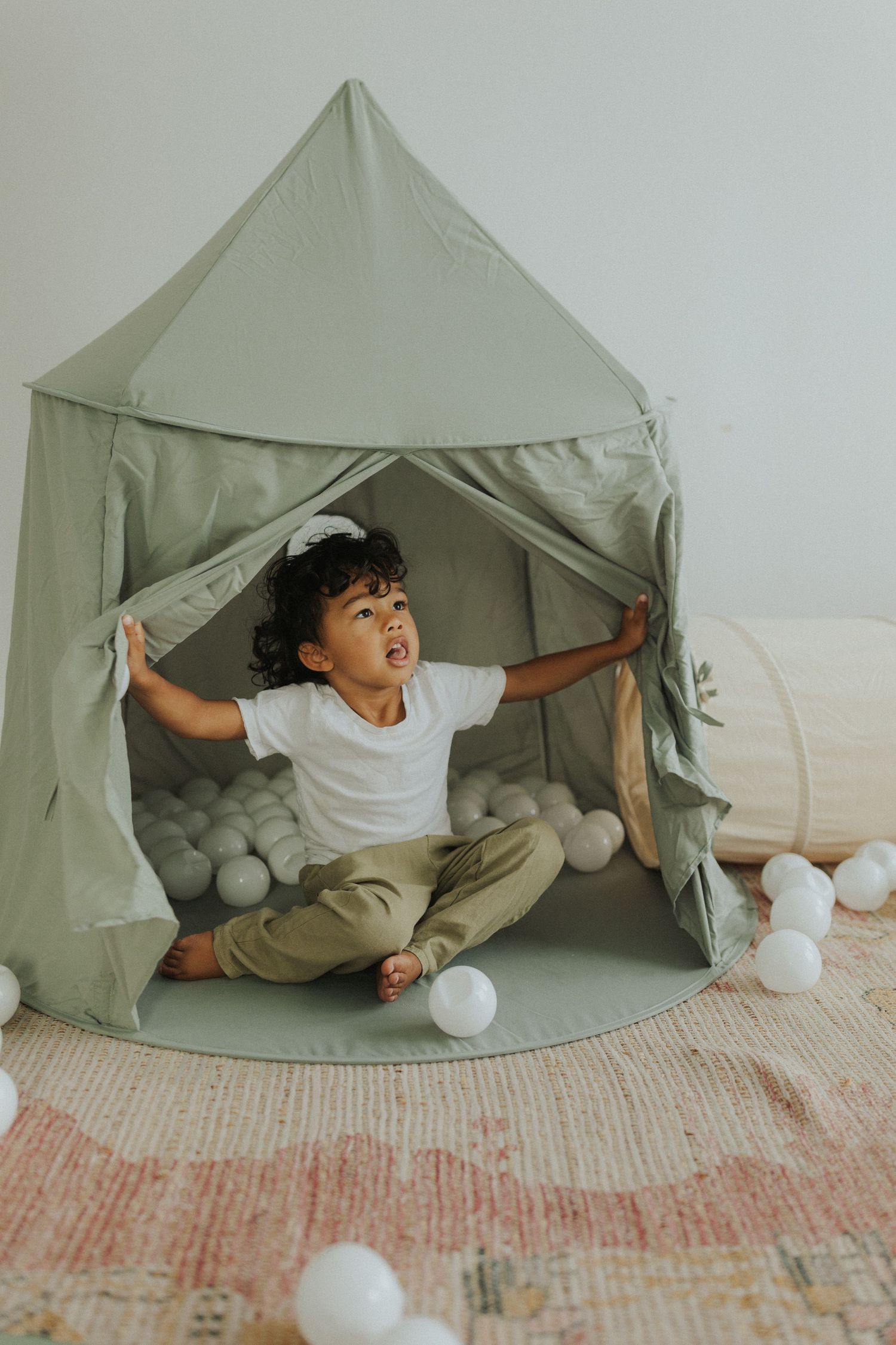 A child plays inside a gray tent fort with white balls scattered around on a textured floor mat.