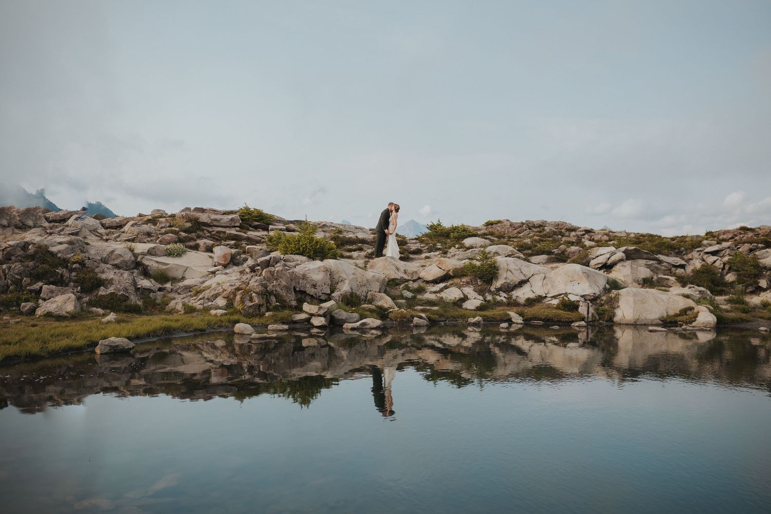 A couple stands on a rocky mountain summit overlooking a misty mountain landscape at sunset.