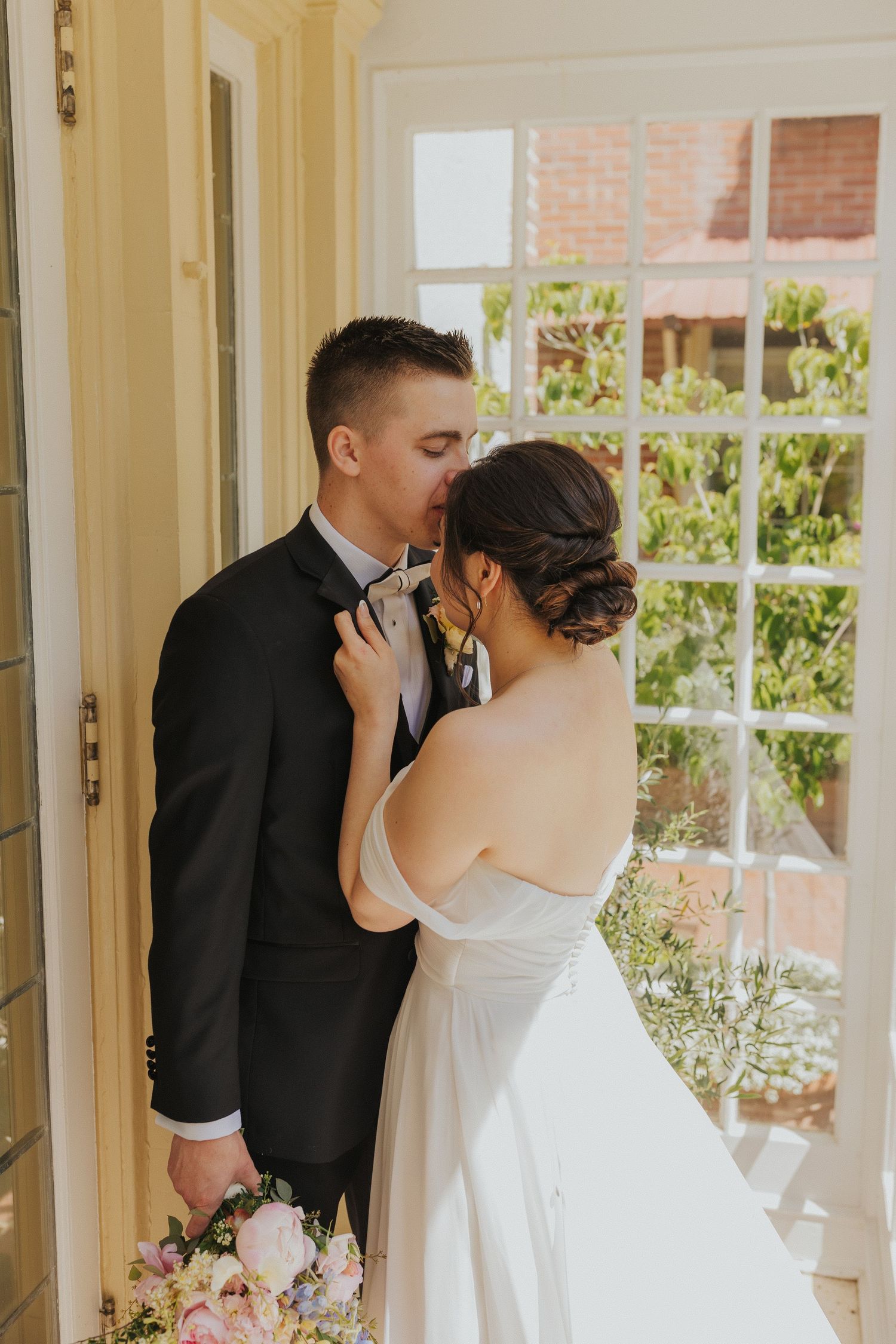 Romantic wedding couple sharing an intimate moment in a sunlit doorway with soft floral details.
