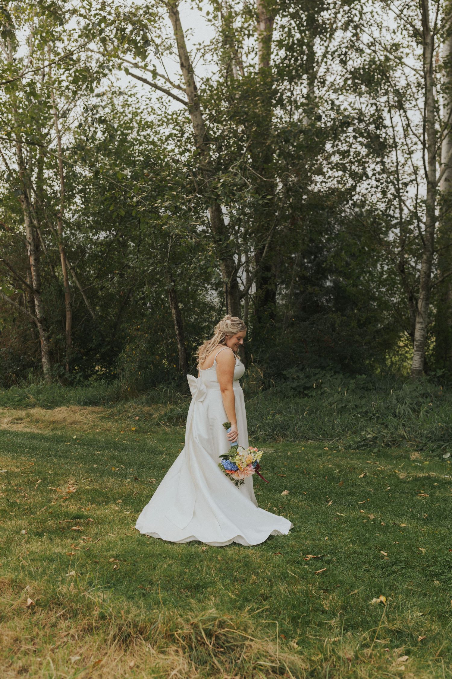 A bride in a white gown twirls gracefully on a grassy hillside surrounded by trees.
