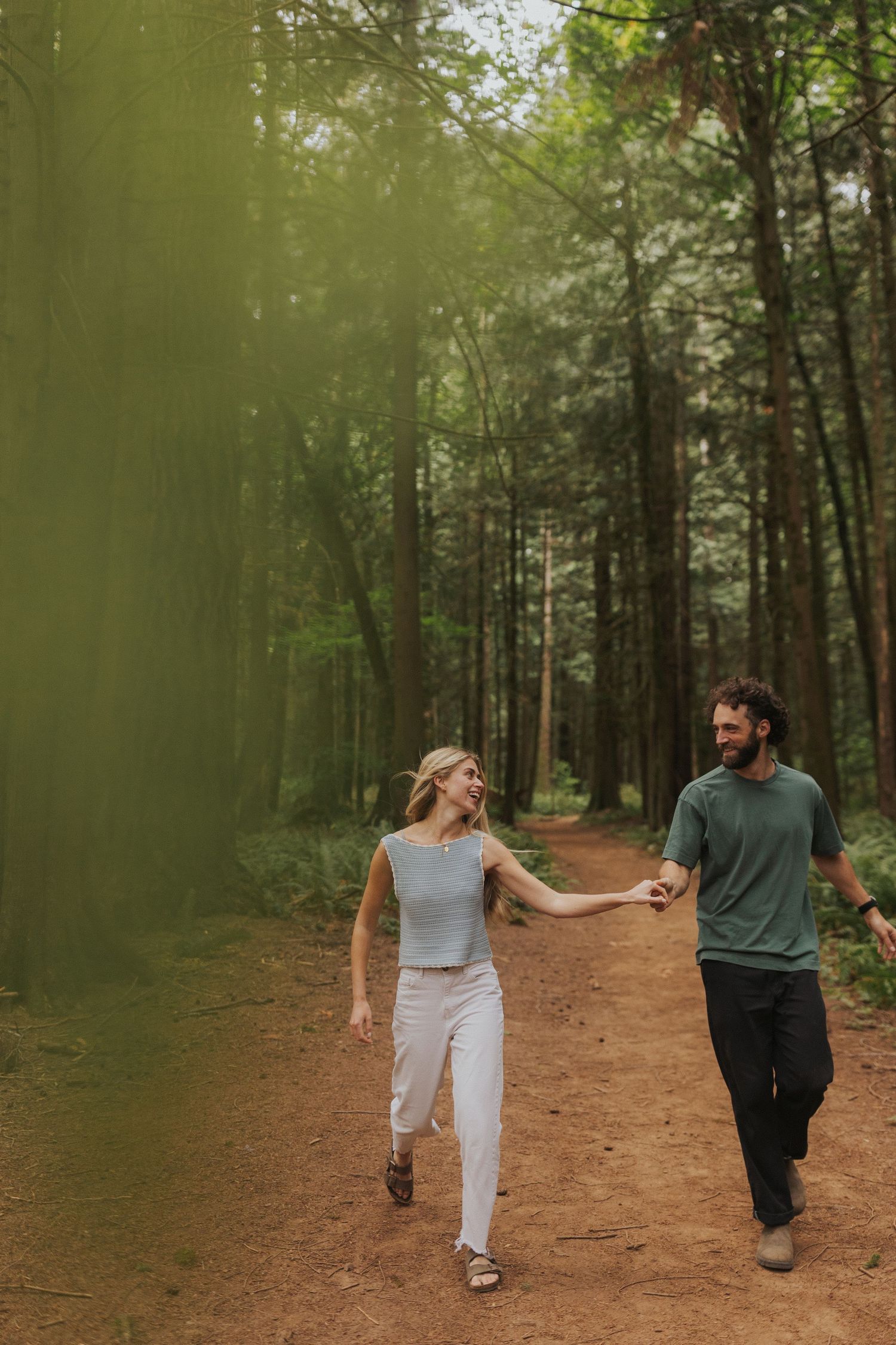 Couple playfully walking and holding hands on a forest trail surrounded by tall trees.