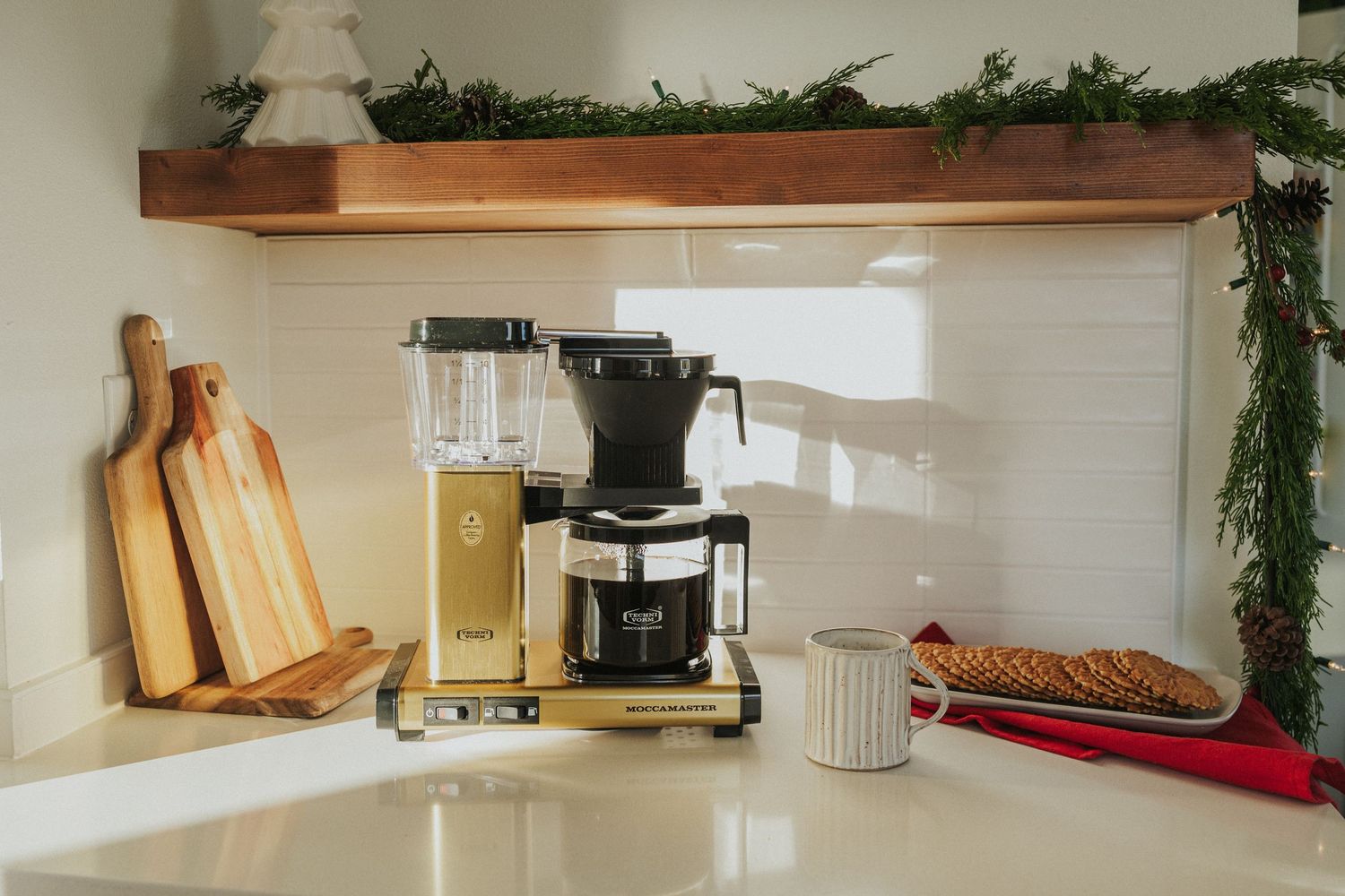 White kitchen counter with coffee maker, cutting boards, and holiday greenery decorations.