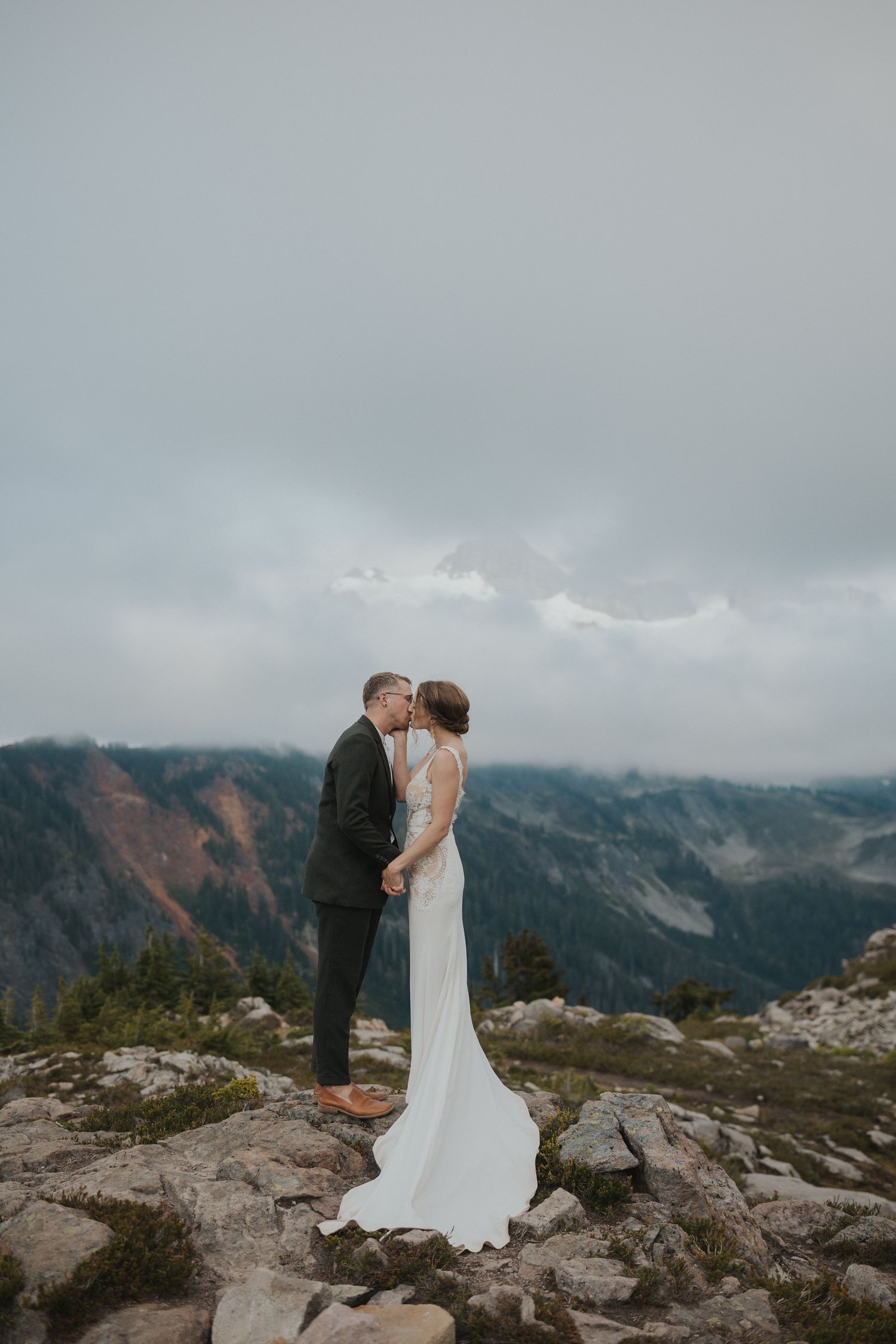 A couple shares an intimate moment on a mountain peak with dramatic cloudy landscape behind them.