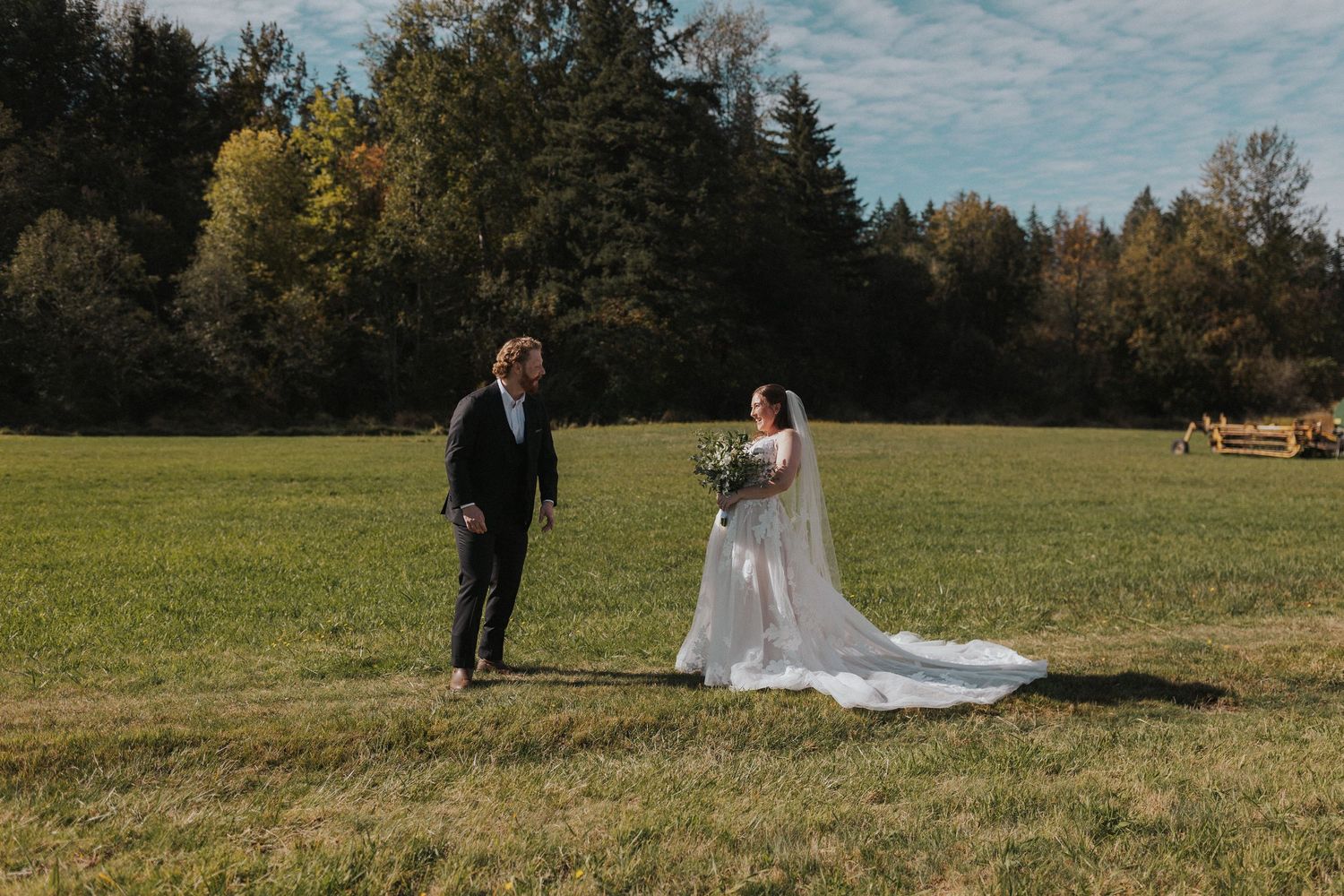 Wedding couple poses together in a sunny meadow with evergreen trees lining the horizon.