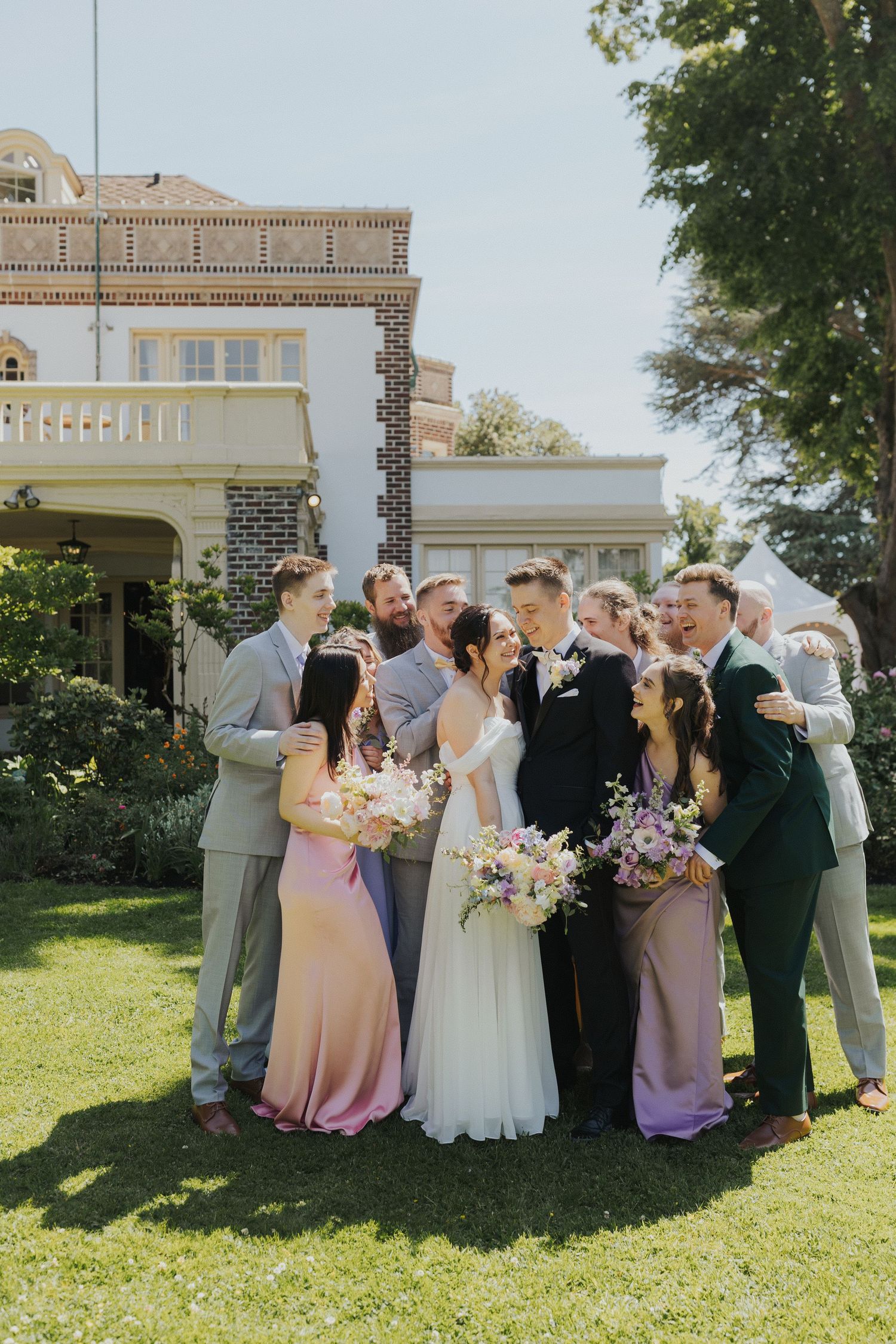 Joyful wedding party gathered in circle on mansion grounds with pastel florals.