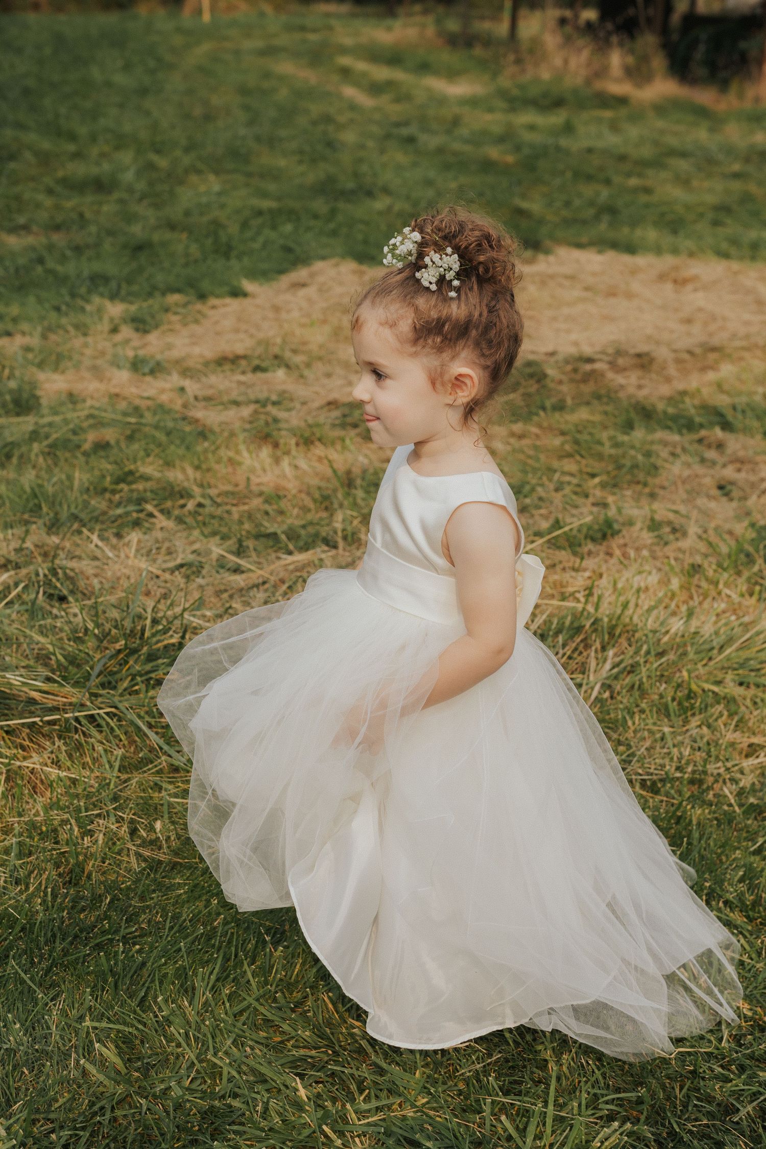 A flower girl in a white tulle dress twirls playfully on the grass during a wedding.