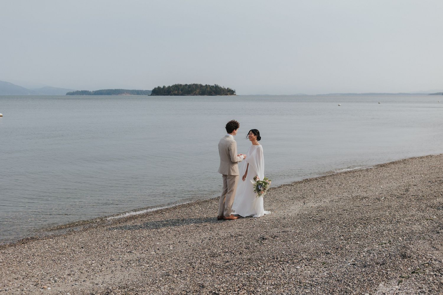 A couple stands together on a peaceful beach shoreline with an island visible in the distance.