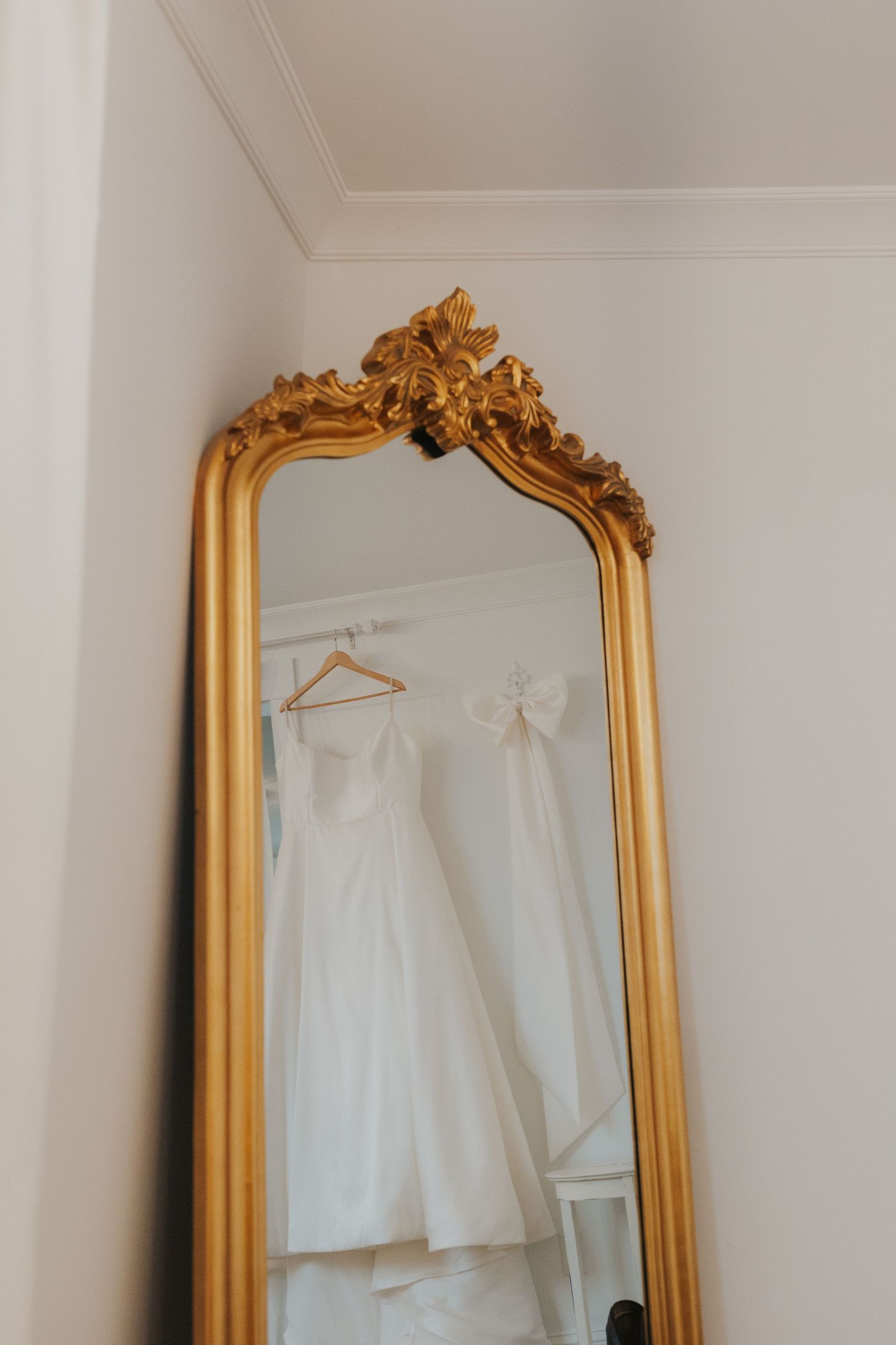 White wedding dress hanging beside an ornate gold-framed mirror in a minimalist room.