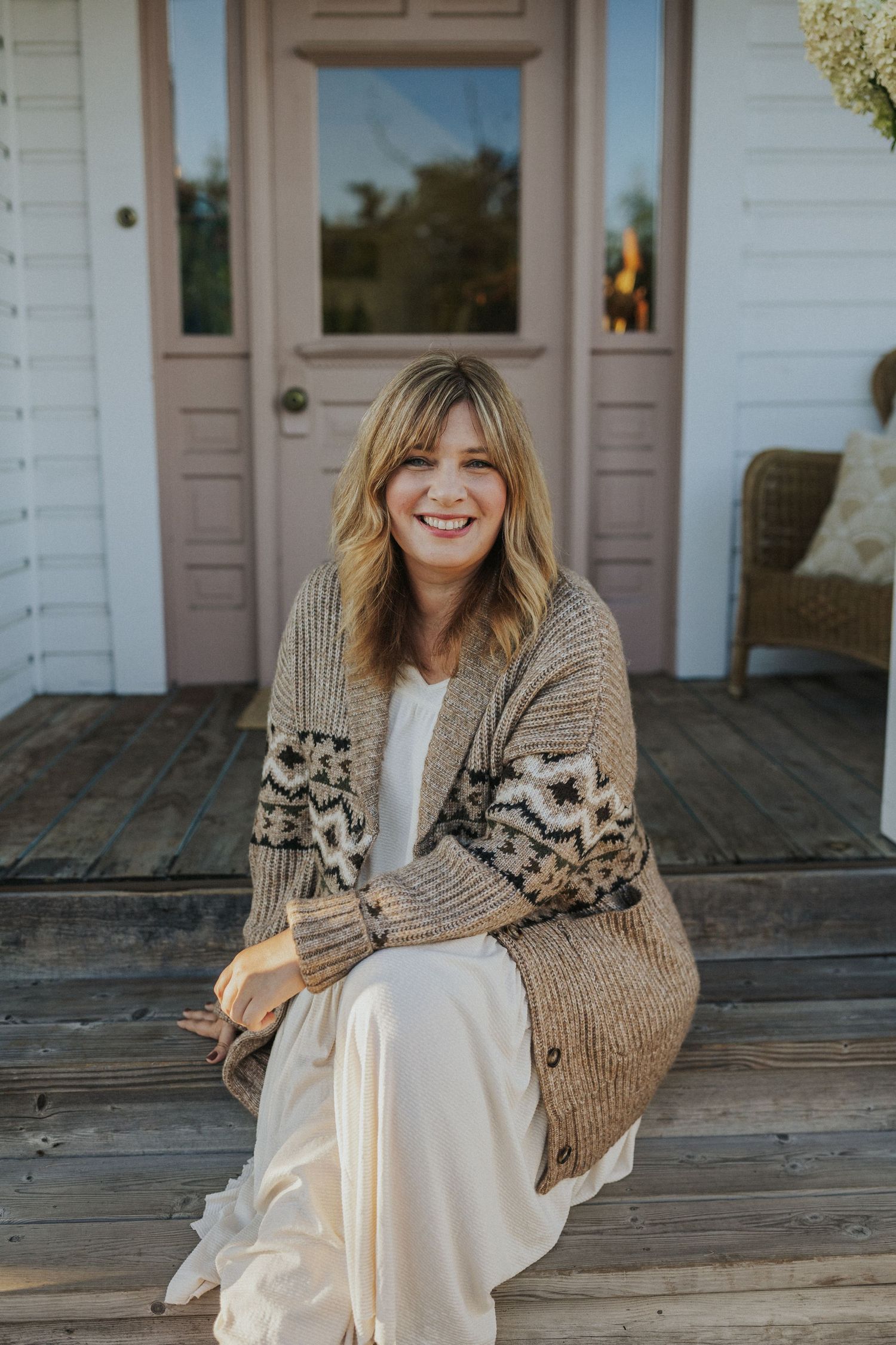 Woman in white dress and cozy knit cardigan sits on wooden porch steps in natural lighting.