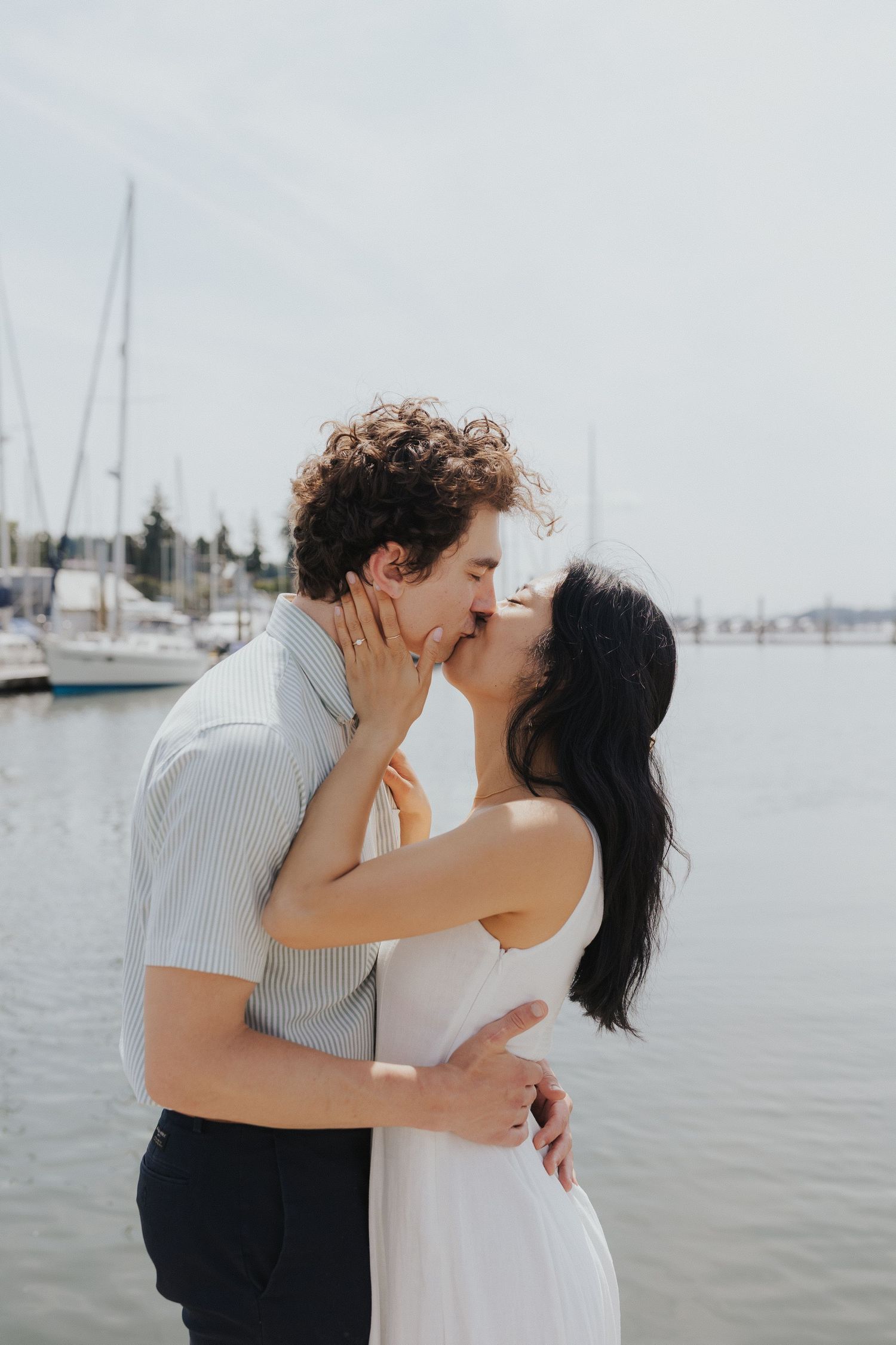 Romantic couple sharing a tender moment by the marina with sailboats in the background.