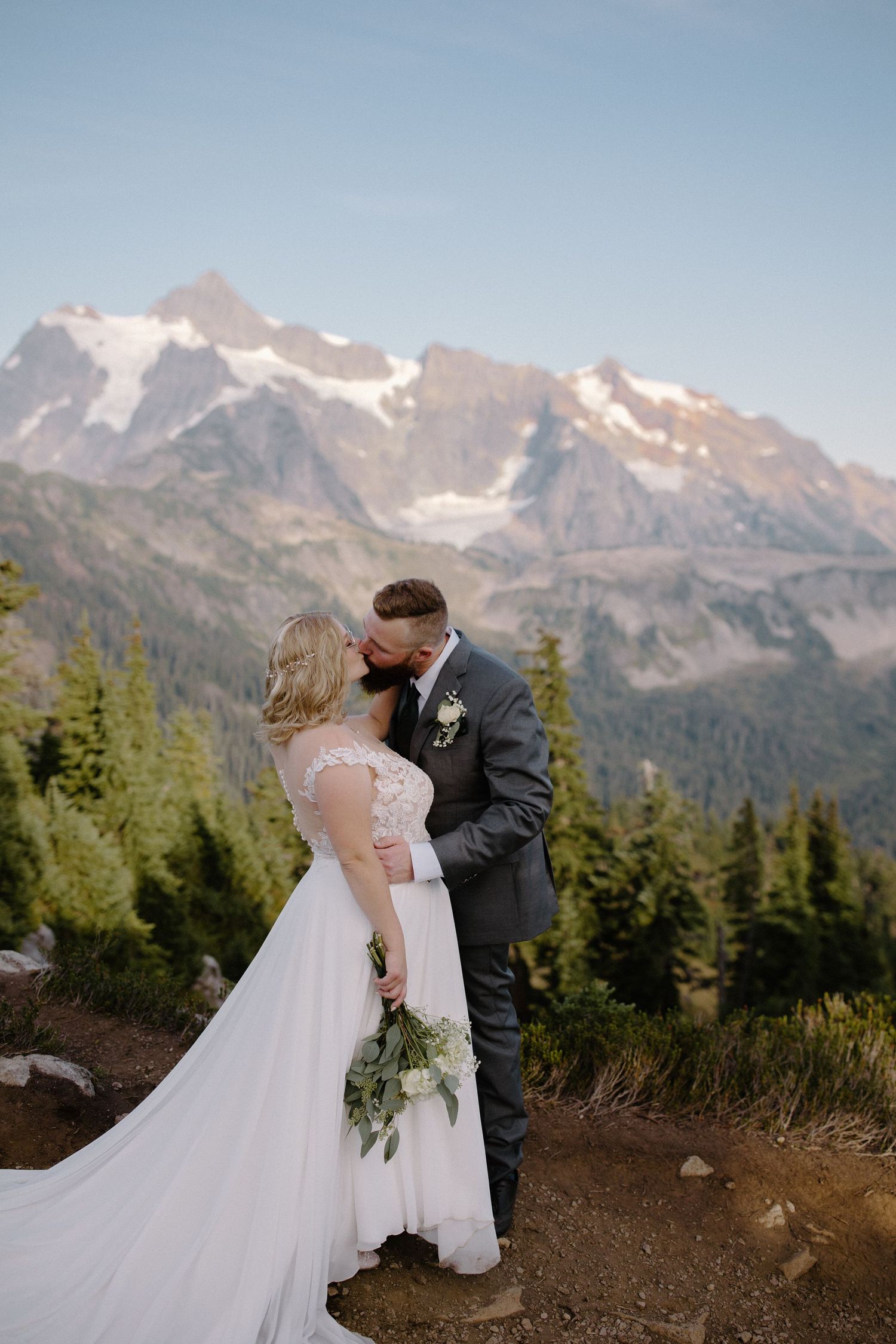 A wedding couple shares an intimate moment against a backdrop of snow-capped mountain peaks and evergreen forest.