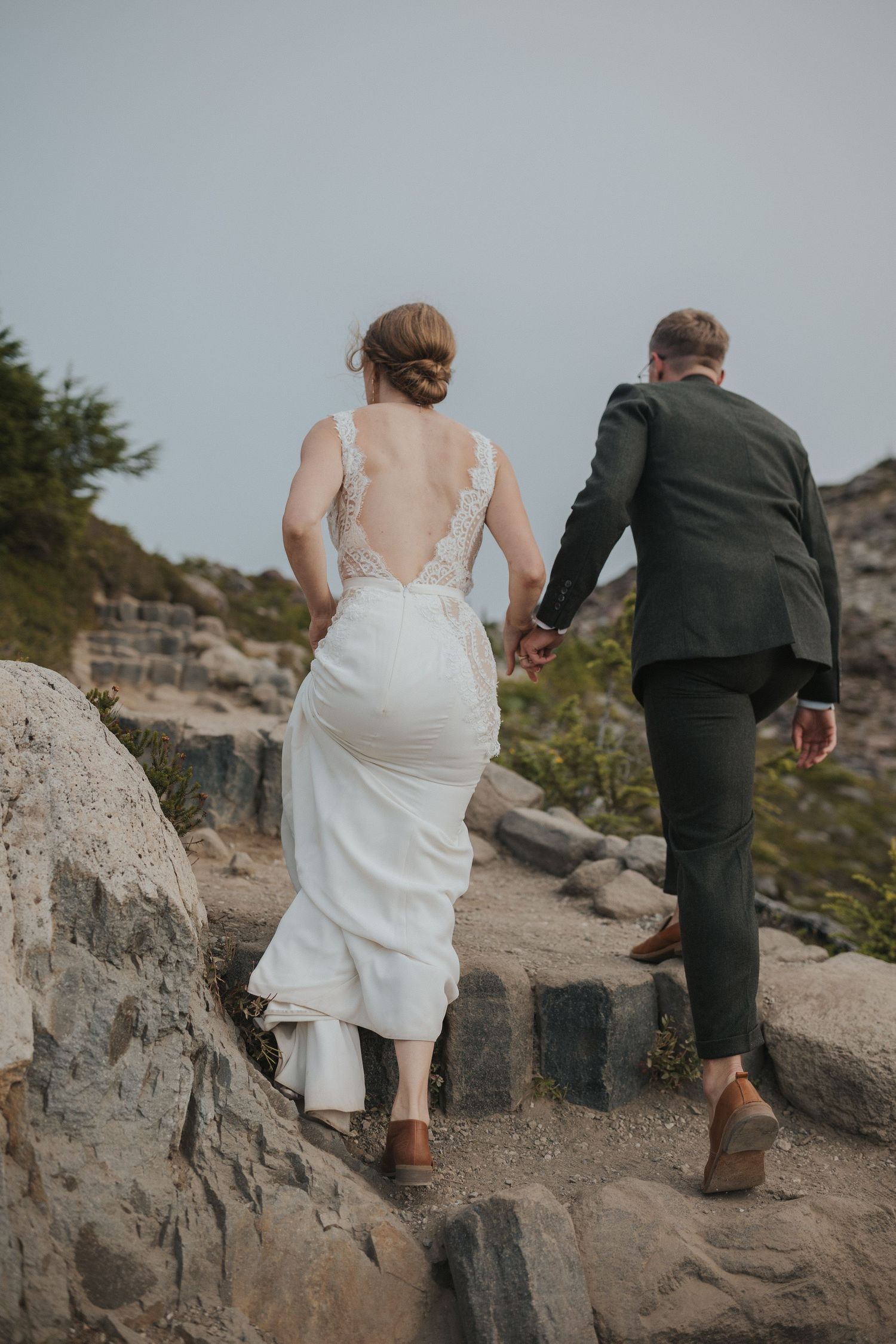 A couple carefully climbs down steep rocky terrain in formal attire.