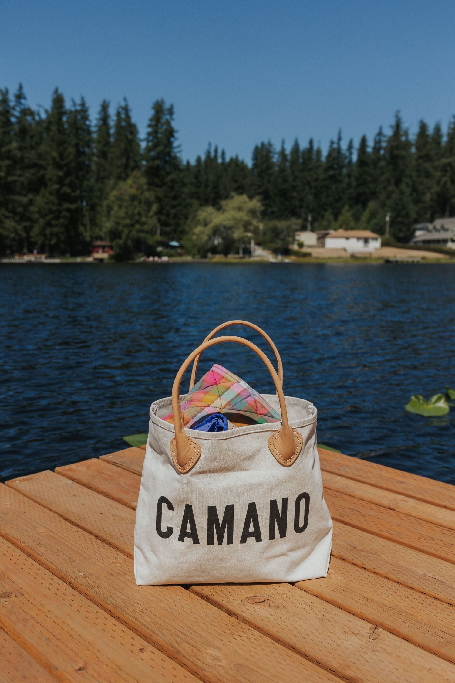 A white canvas Camano bag sits on wooden dock planks with blue water and trees in background.