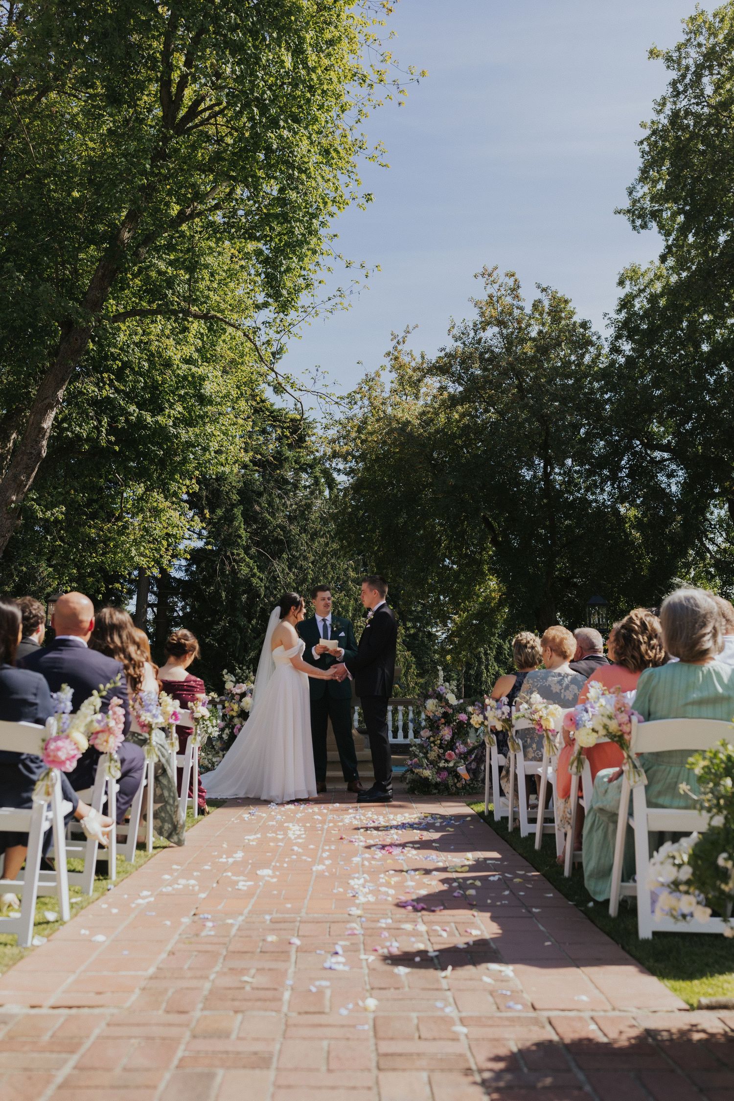 Outdoor wedding ceremony setup with white chairs and rose petals lining brick pathway.