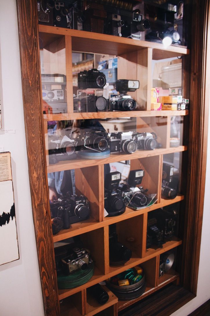 Vintage camera collection displayed in wooden shelving unit behind glass.