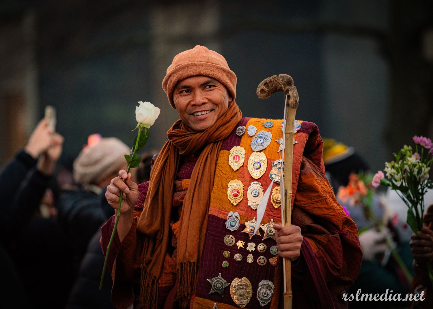 Person in orange winter clothing holds white flower and displays sash decorated with various pins and badges at outdoor event.