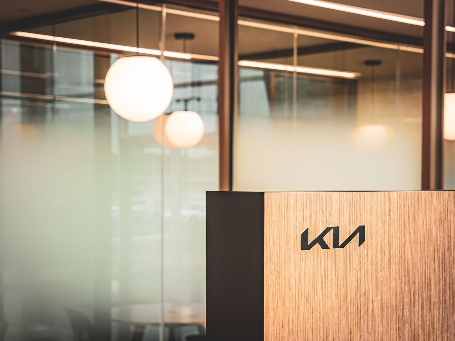 Modern office interior with glass walls, pendant lighting, and a KIA logo sign mounted on a wood-paneled wall.