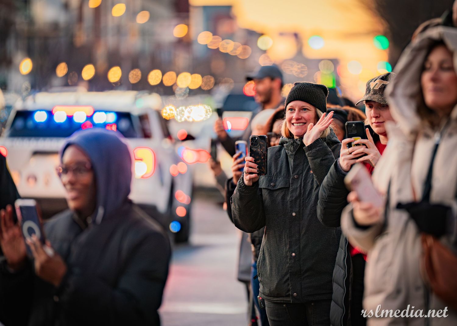 A crowd of people watching and photographing an event at dusk with bokeh lights in the background.