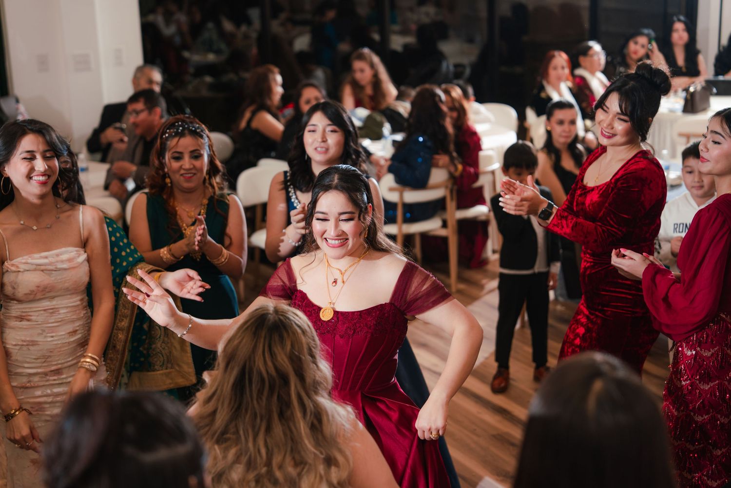 Wedding guests dancing and celebrating on a wooden dance floor at an indoor reception venue.