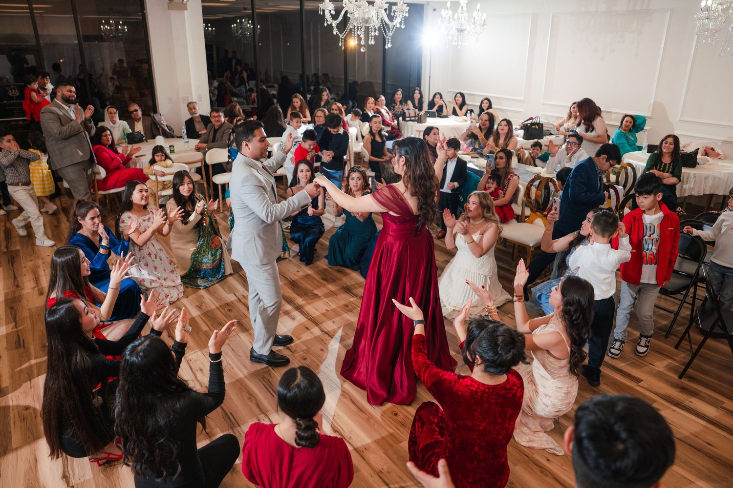 Wedding guests dance and celebrate together on a wooden dance floor in an elegant ballroom with chandeliers.