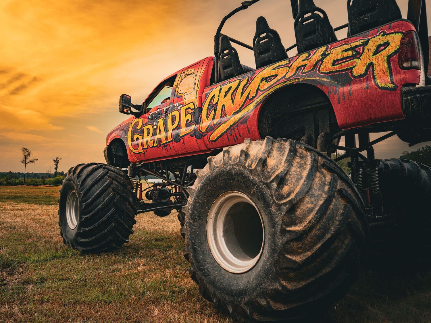 Monster truck silhouetted against dramatic orange sunset during outdoor motorsports event.