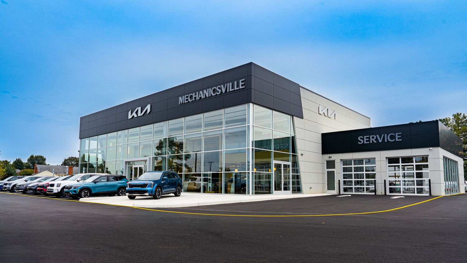 A panoramic exterior view of a modern Kia car dealership with glass walls and new vehicles on display in the lot.