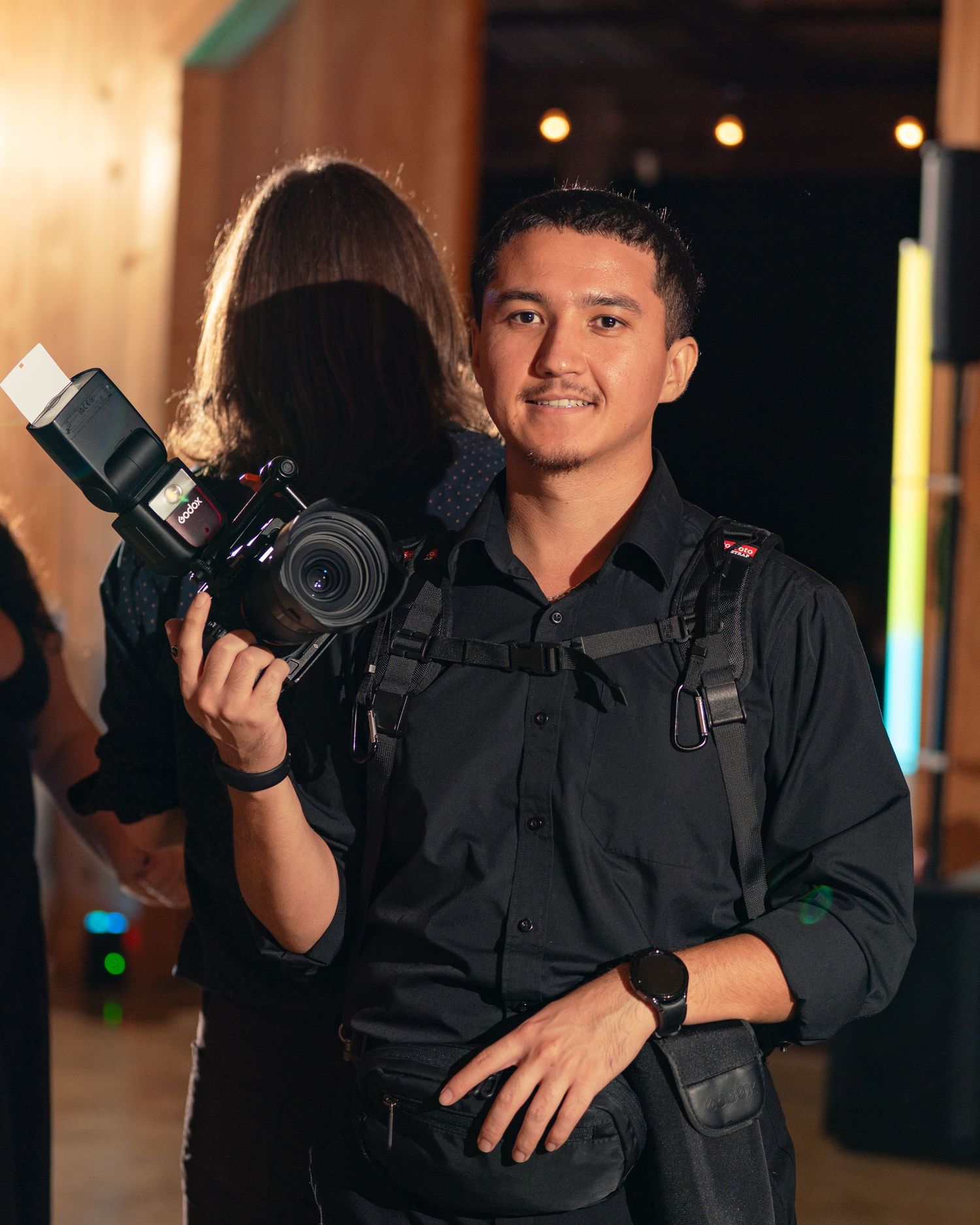 Photographer in a black shirt holds a professional camera while wearing a harness and smartwatch at an indoor event with warm lighting.
