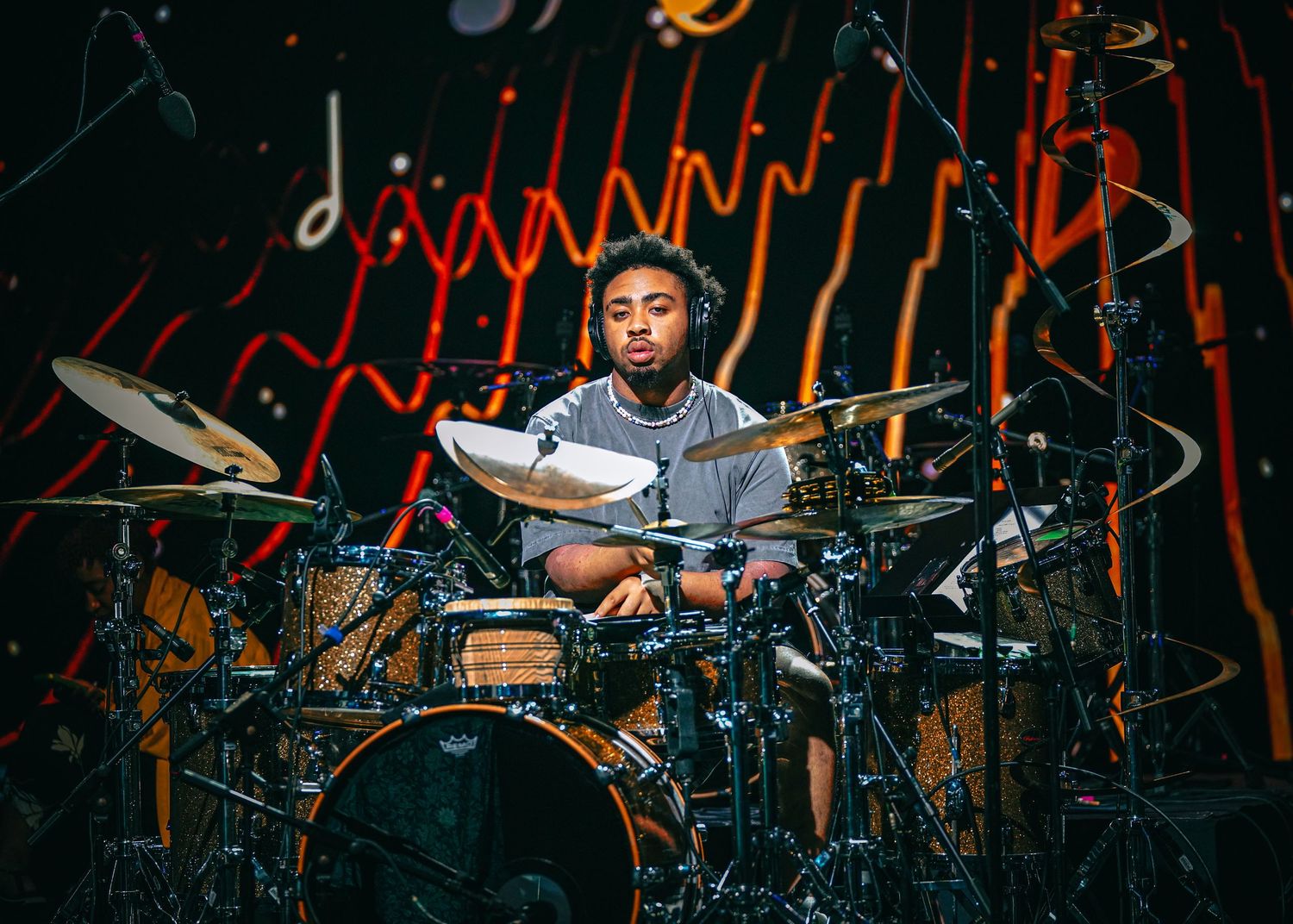 Drummer performing on stage with dynamic lighting and multiple drum cymbals visible in the dark atmosphere.