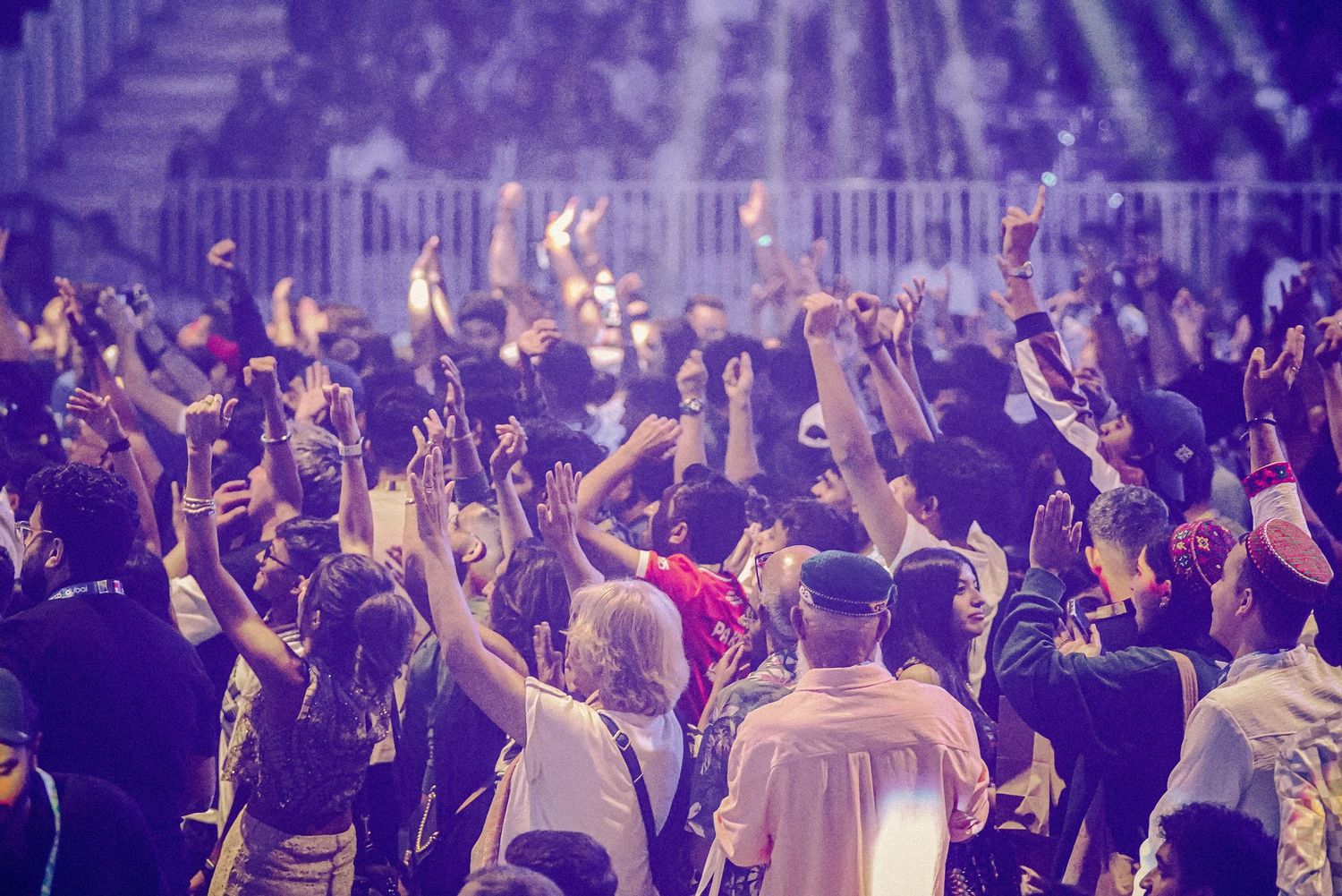 Concert crowd raises hands in a dark arena illuminated by purple stage lights during live performance.