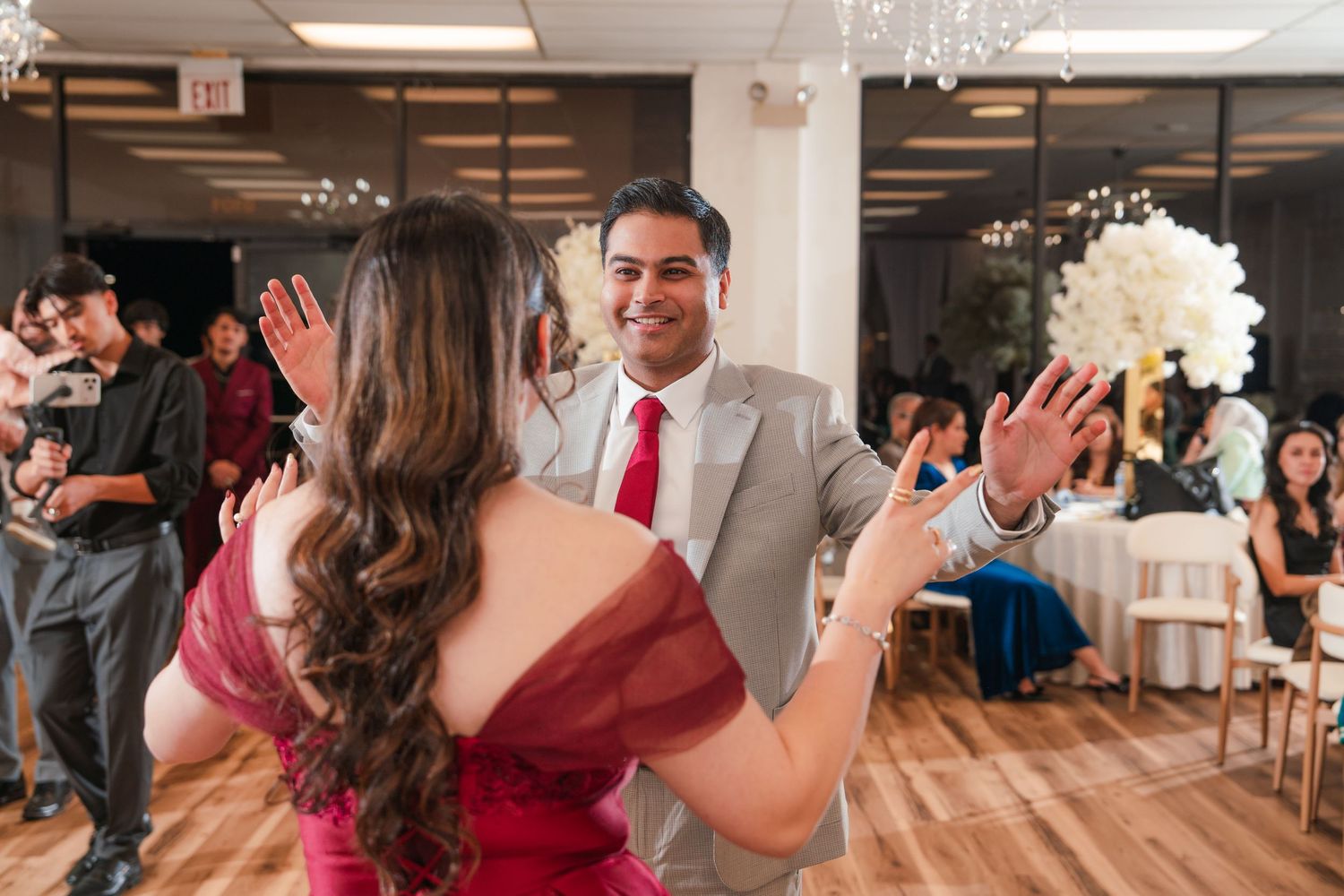 Wedding guests dancing and celebrating on a dance floor at an indoor reception venue with wooden accents and chandeliers.
