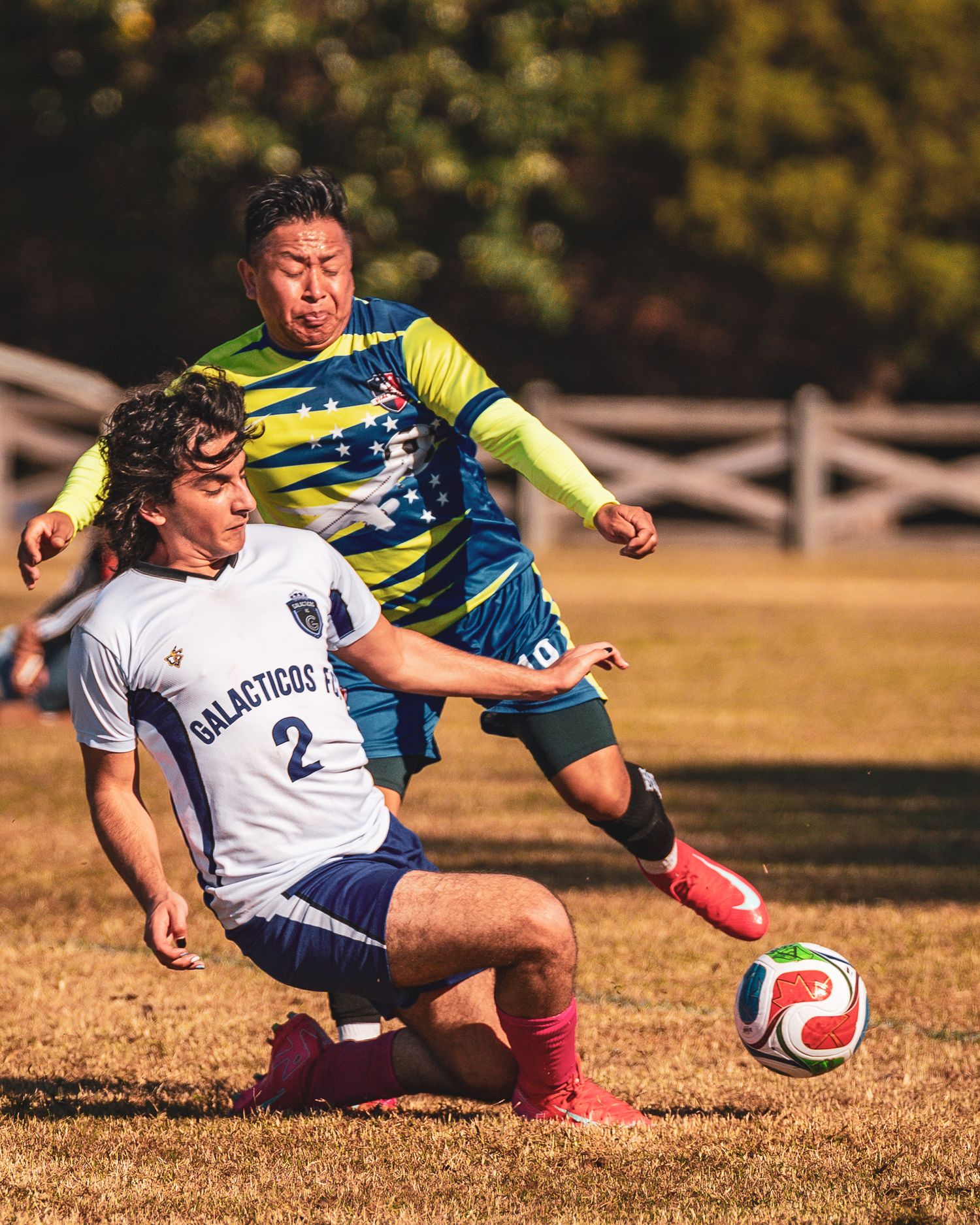 Soccer players collide while competing for the ball during an intense match on a dry grass field.