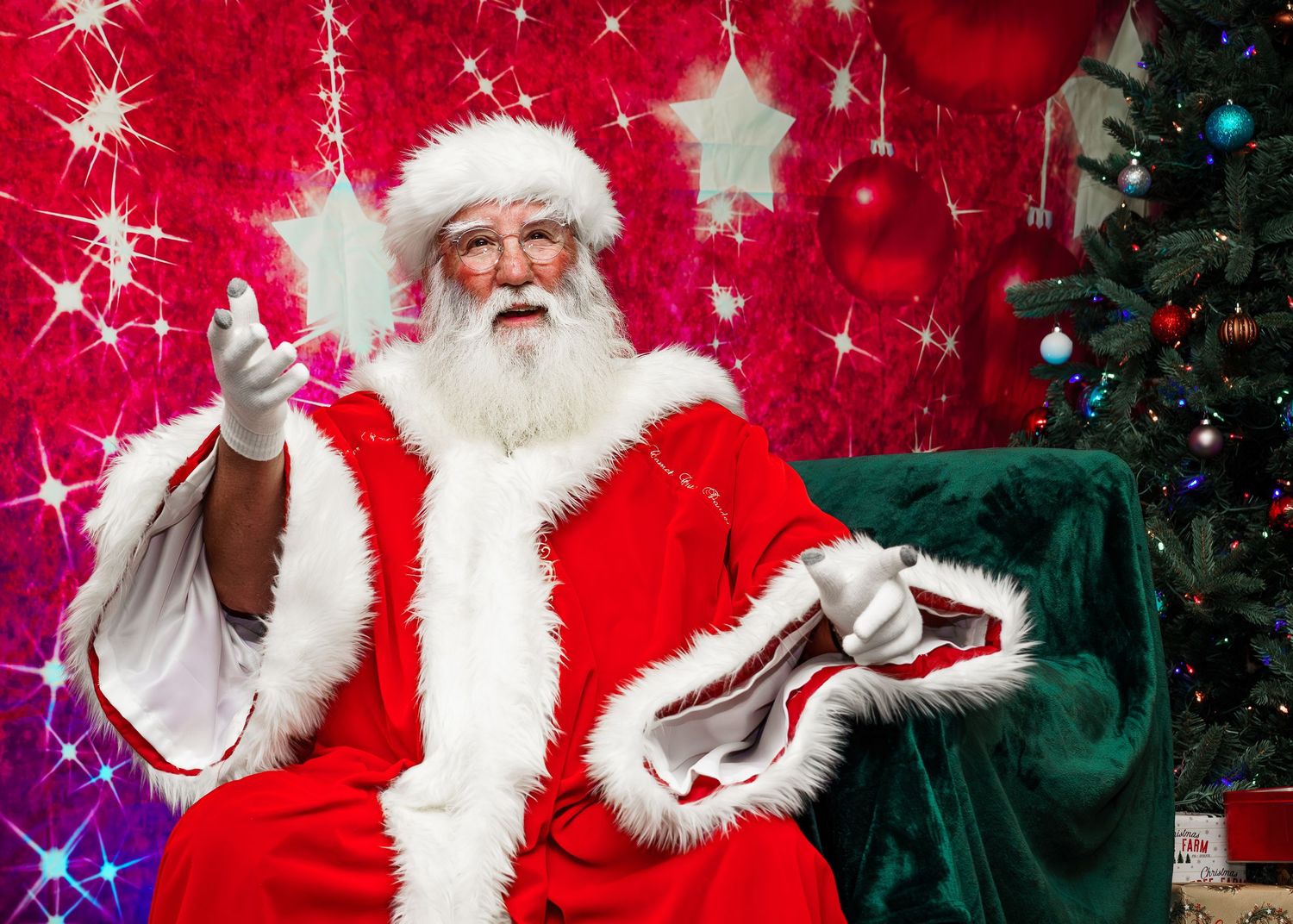 Traditional Santa Claus figure in red suit gestures while sitting next to decorated Christmas tree with sparkling red background.