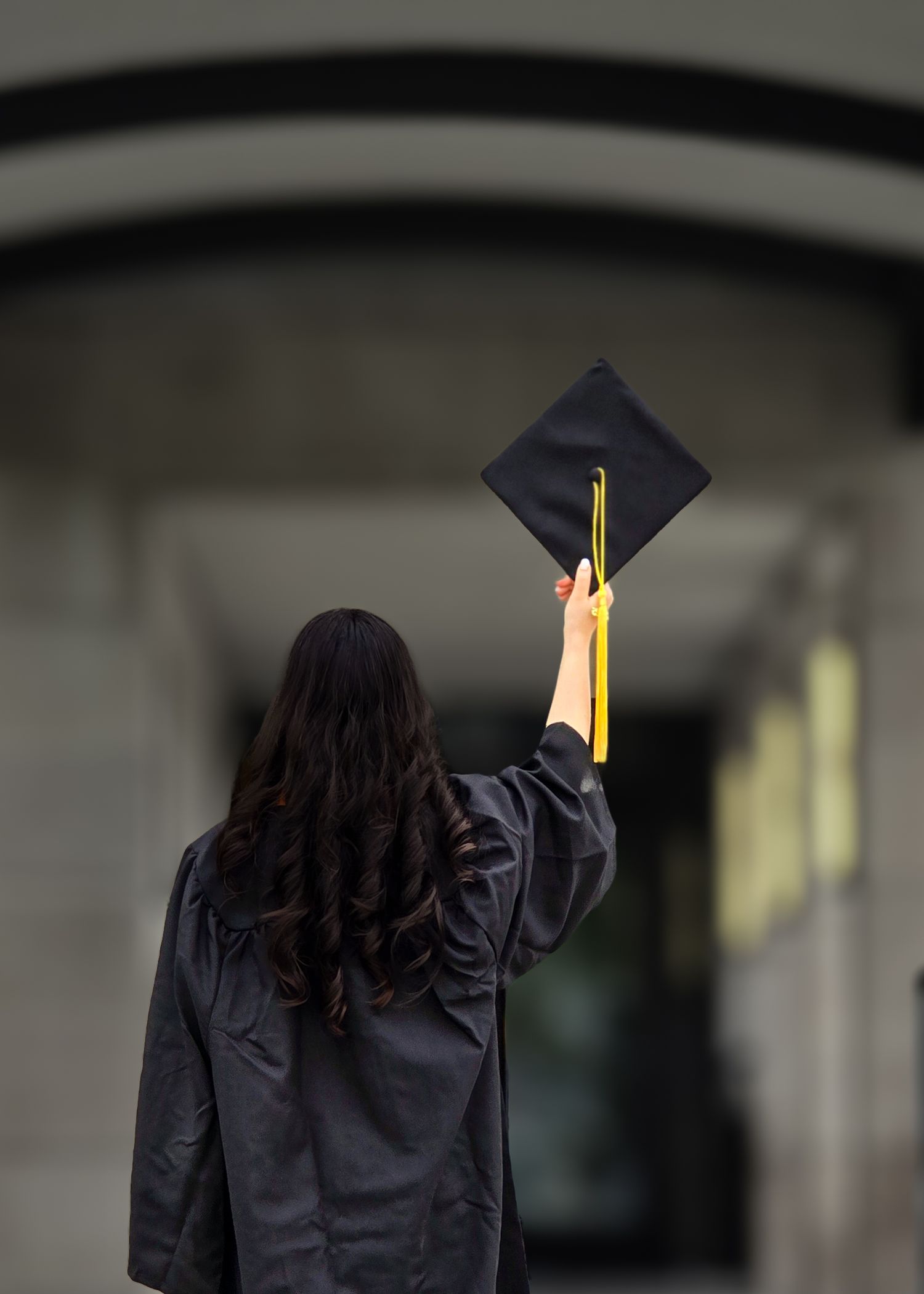 Graduate in black cap and gown holds up their mortarboard cap from behind.
