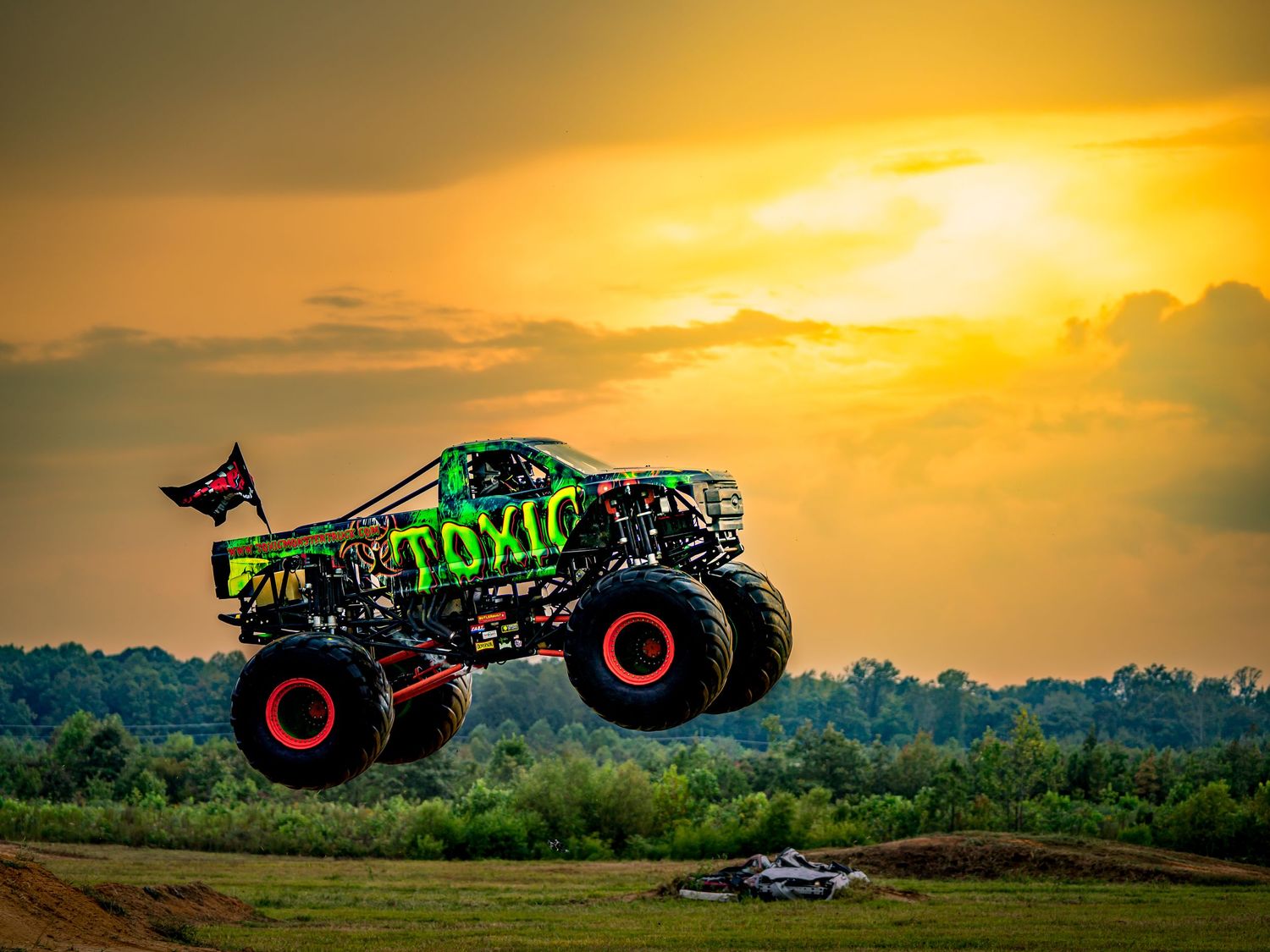Green monster truck with Toxic branding launches off dirt ramp at sunset.