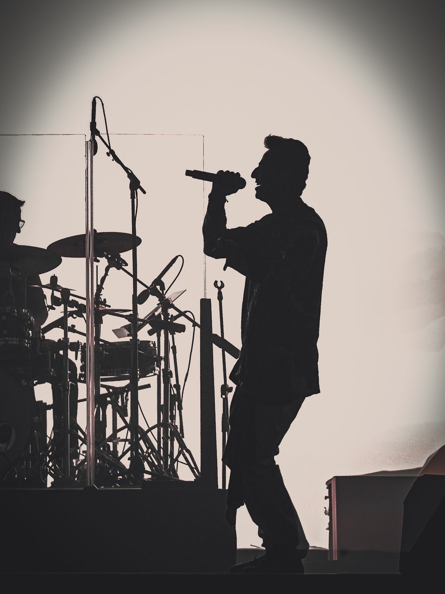 Silhouette of a singer performing on stage with a microphone and drum kit visible in dramatic backlit concert lighting.