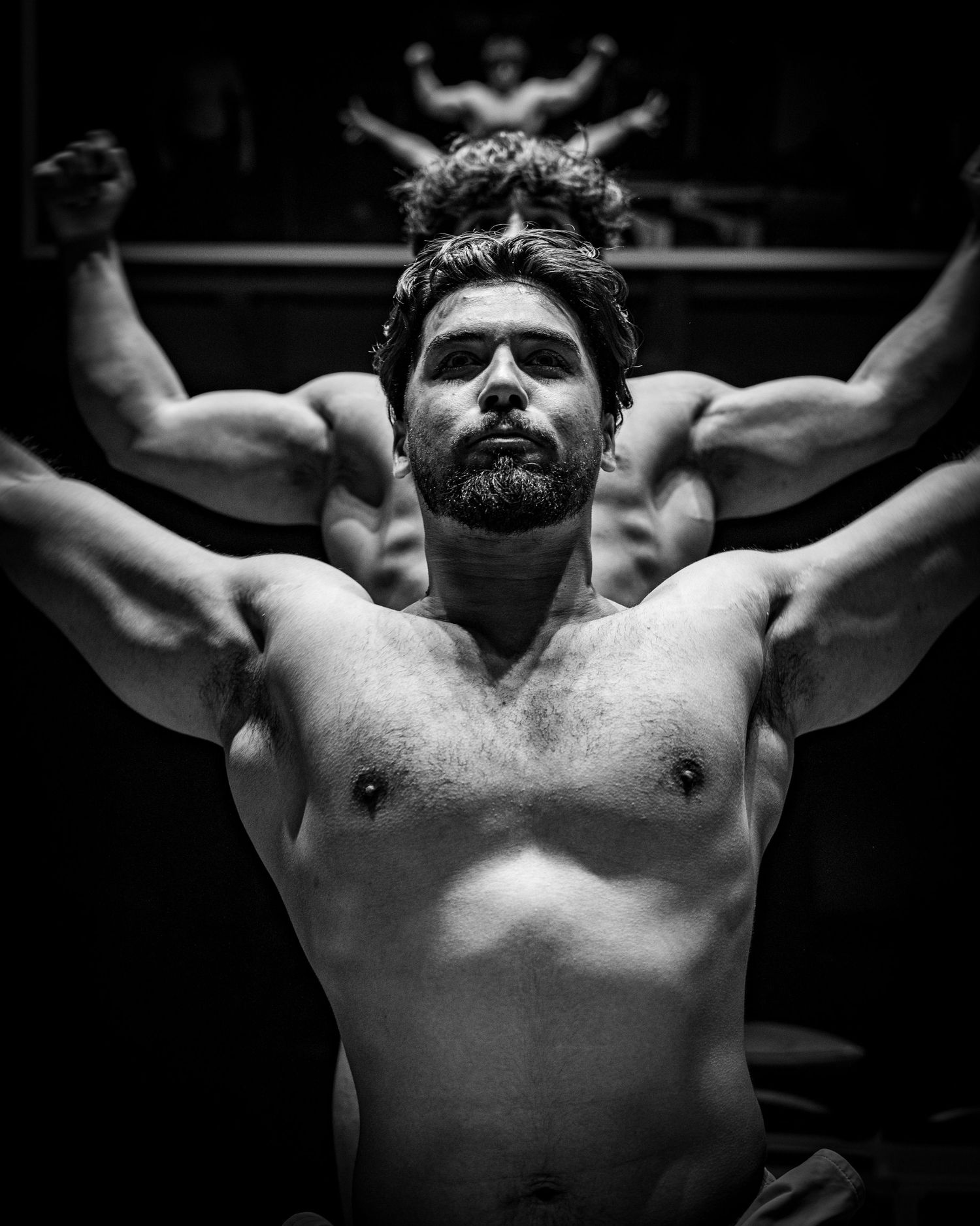 Black and white artistic portrait of a muscular shirtless athlete with arms raised in dramatic lighting.