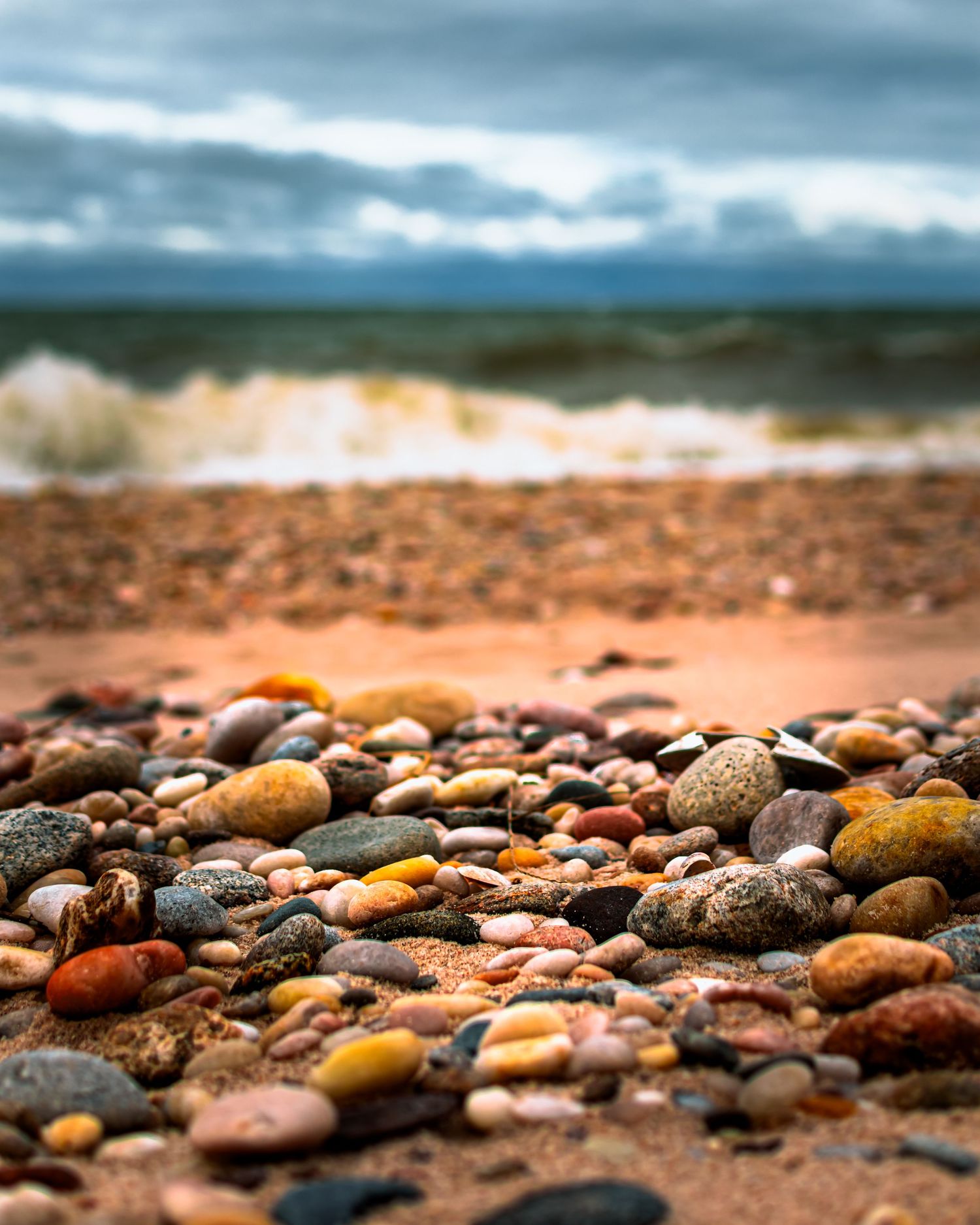 Colorful pebbles and stones scattered along a beach shoreline with dramatic cloudy sky in the background.