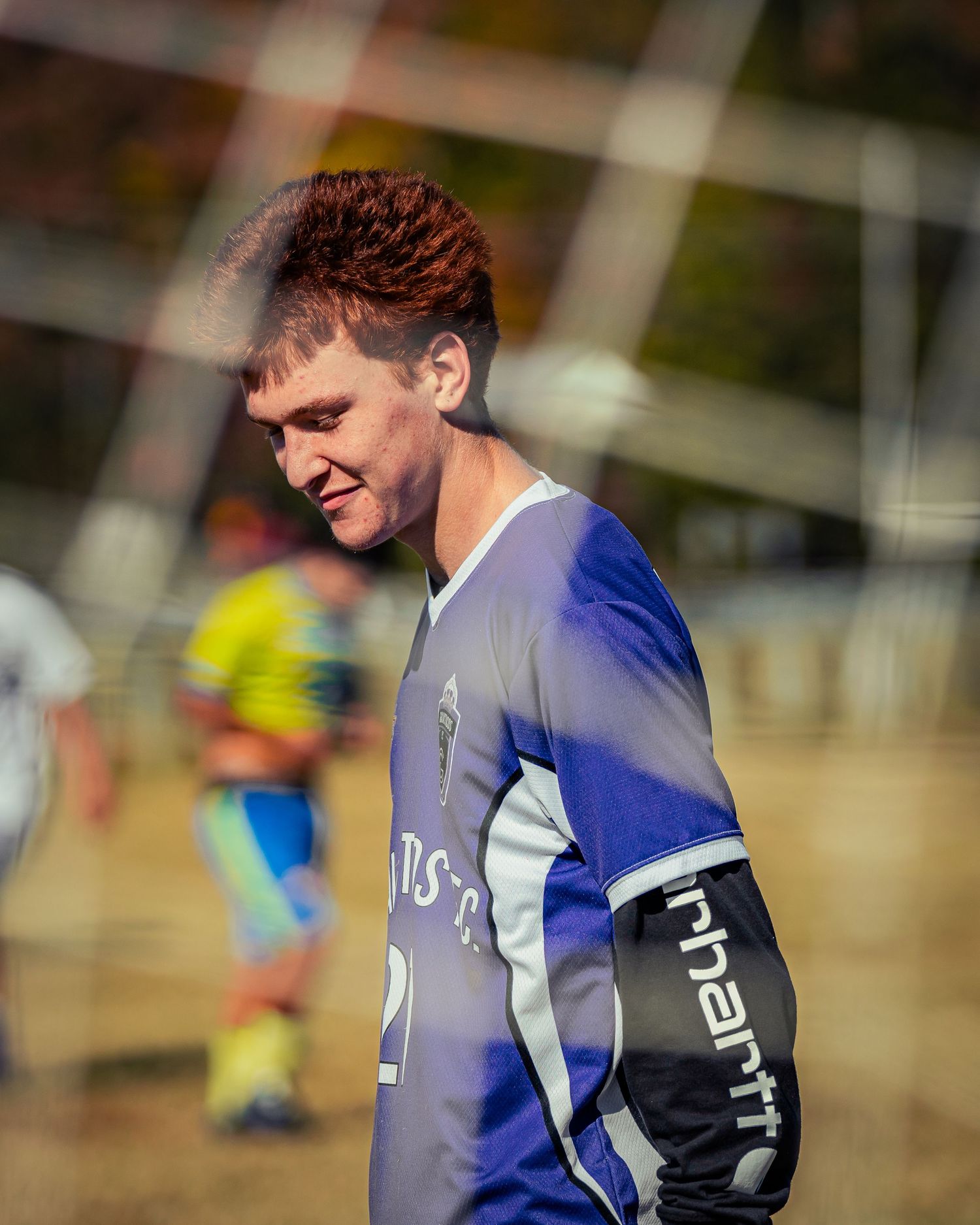 Soccer player in blue and white jersey with black arm sleeve stands on field during outdoor match in sunlight.