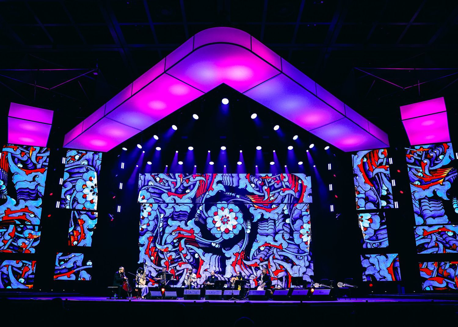 A concert stage illuminated with vibrant purple lighting and colorful LED screens displaying abstract patterns.