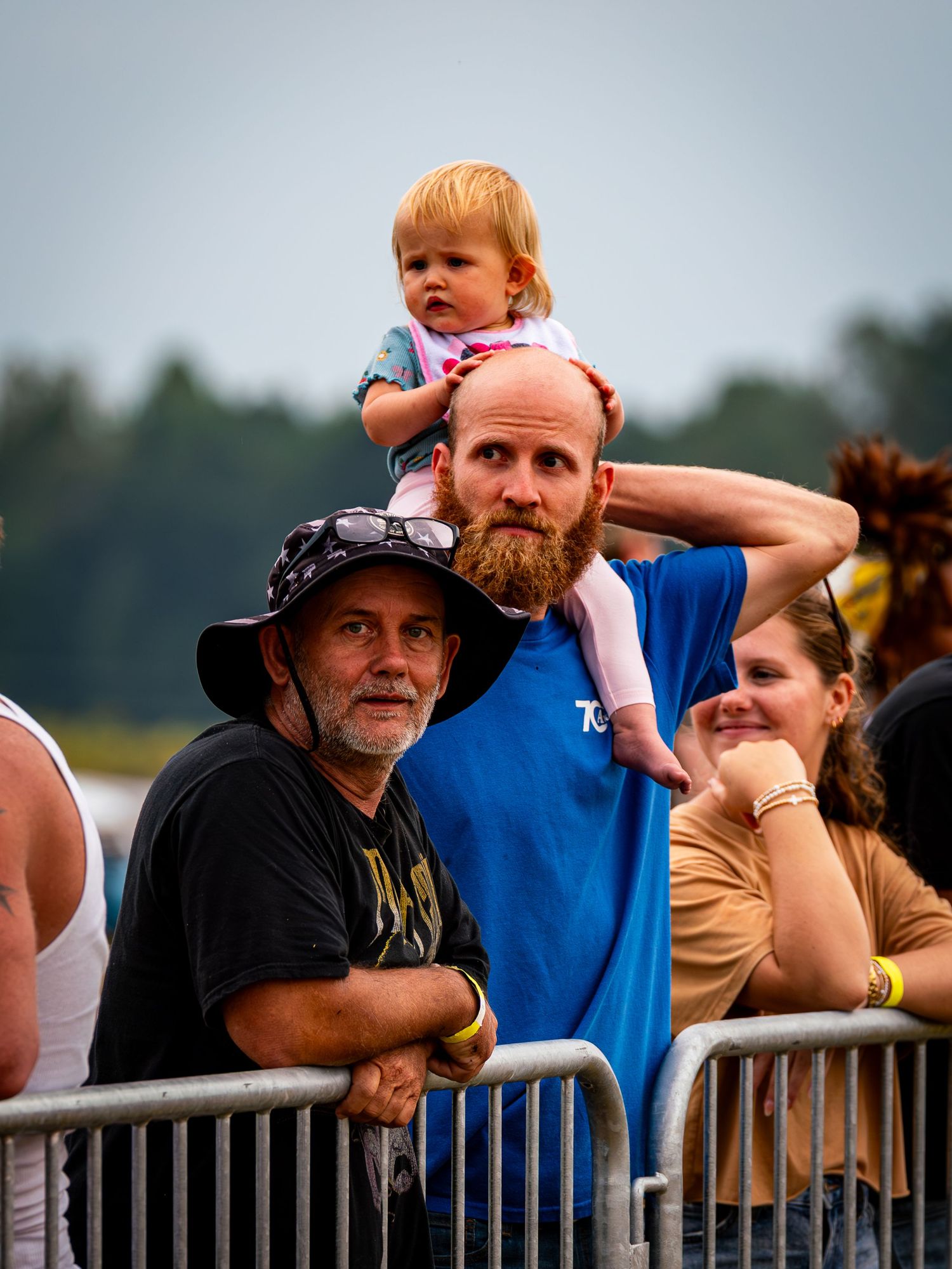 Spectators lean against a metal railing at an outdoor event with a small child visible in the background.