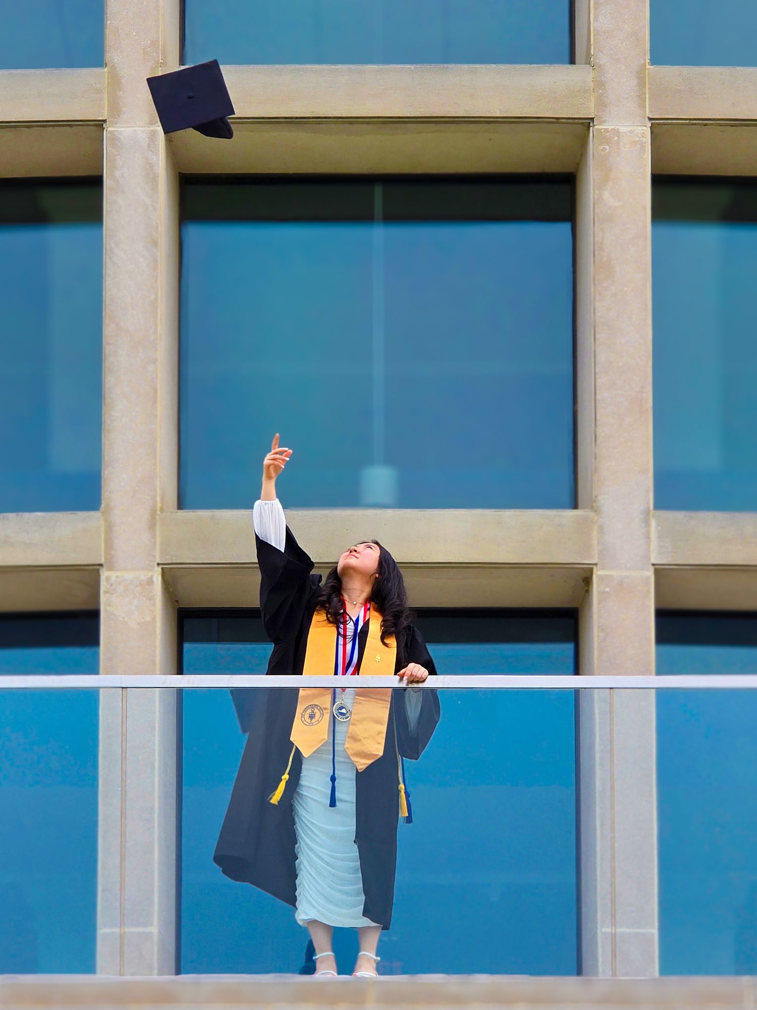 Graduate in regalia stands against modern building while tossing cap in celebration.